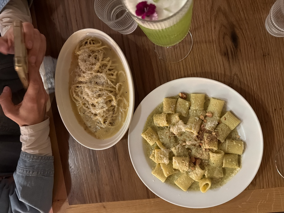 Two pasta dishes sit on a wooden table. One, a long, thin pasta covered in a creamy sauce and shredded cheese, is served in an oblong white bowl. The other features a larger, tube-shaped pasta in a green sauce, also topped with cheese and chopped nuts, presented on a round white plate. A green drink garnished with a flower sits nearby. A person's arm and hand holding a phone are visible to the left of the oblong pasta dish.