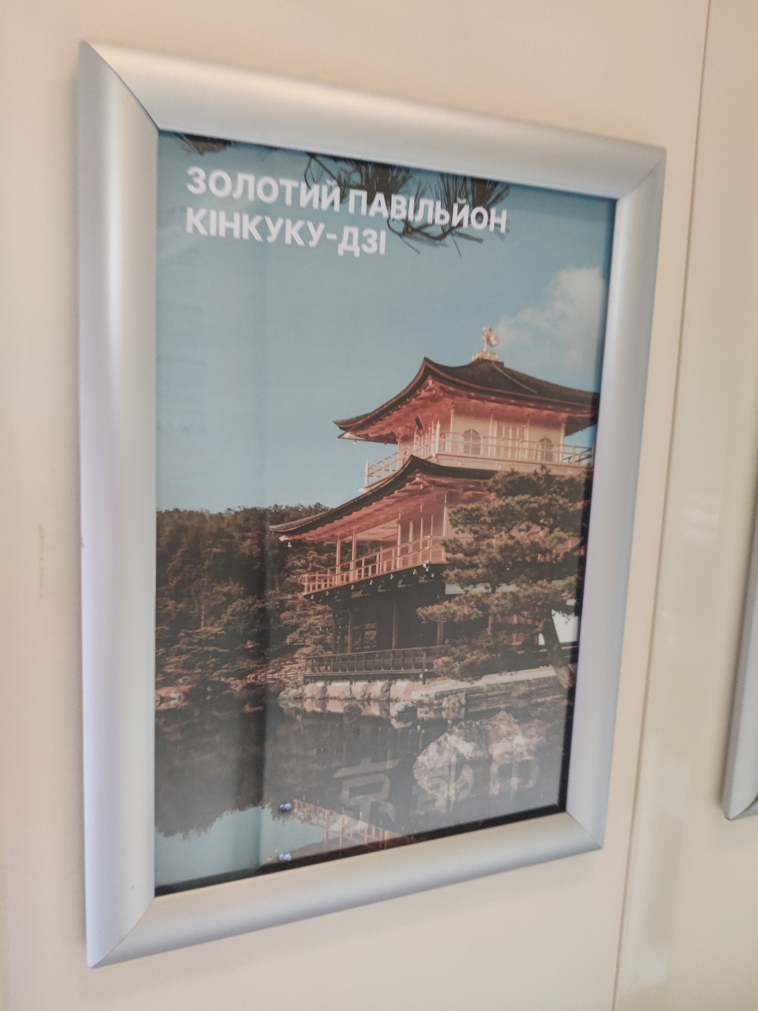 A framed photograph of the Kinkaku-ji (Golden Pavilion) temple in Kyoto, Japan, is displayed on a wall. The photograph shows the two-story Zen Buddhist temple, covered in gold leaf, reflecting in the surrounding pond. Lush trees and foliage are visible around the temple. White text in a Cyrillic alphabet is printed at the top of the photograph, which reads "ЗОЛОТИИ ПАВИЛЬОН КІНКУКУ-ДЗІ" (Golden Pavilion Kinkaku-ji). The frame is silver and appears to be made of metal. The wall on which the photograph is hung is a light, neutral color. The image is taken from a slightly angled perspective. The text suggests this photograph may be part of an exhibition or display related to tourism or culture, possibly in Sviatopetrivske, Ukraine, or a location related to Ukrainian interest in Japanese culture.