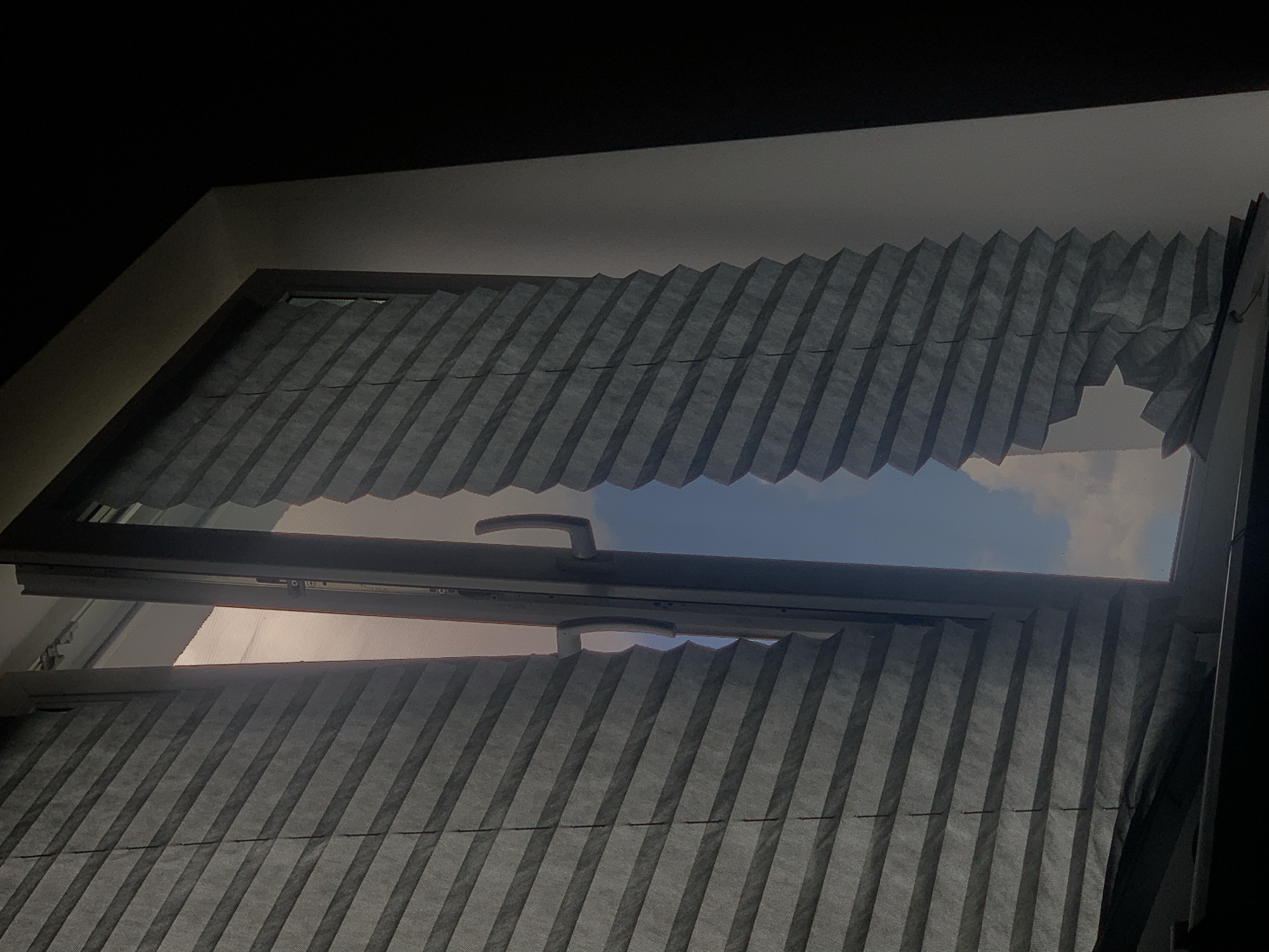 The image depicts a window with grey, pleated blinds, partially open to reveal a view of the sky and clouds. The window frame is dark, and the blinds appear to be made of a fabric material. The scene is dimly lit, with the exterior light creating a contrast with the darker interior. There are no people or animals visible in the image. The location is not specified in the image or provided in the prompt.