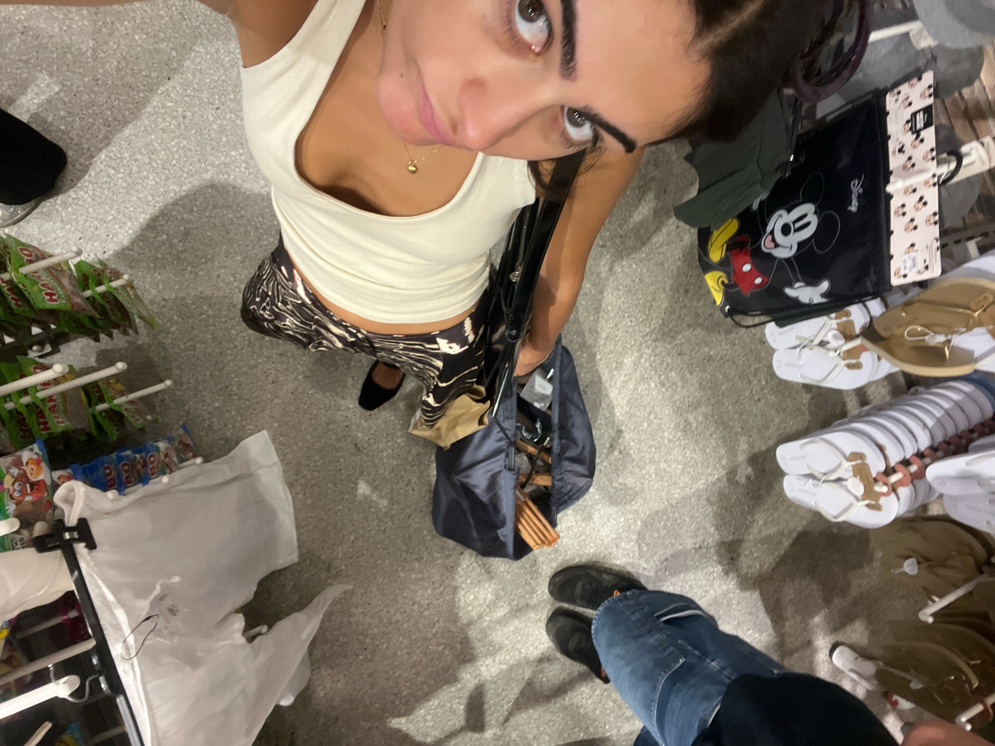 The image shows a high-angle shot of a young woman in a store, likely in Madrid, Spain. She is wearing a white tank top and patterned pants, holding a black bag. Her right foot is visible, wearing a dark shoe. In the background, shelves are stocked with various items, including bags with Mickey Mouse designs and a display of white flip-flops. A man's legs and feet, wearing blue jeans and dark shoes, are visible in the lower right corner, suggesting he is accompanying the woman or is another shopper. The floor is a speckled gray surface.