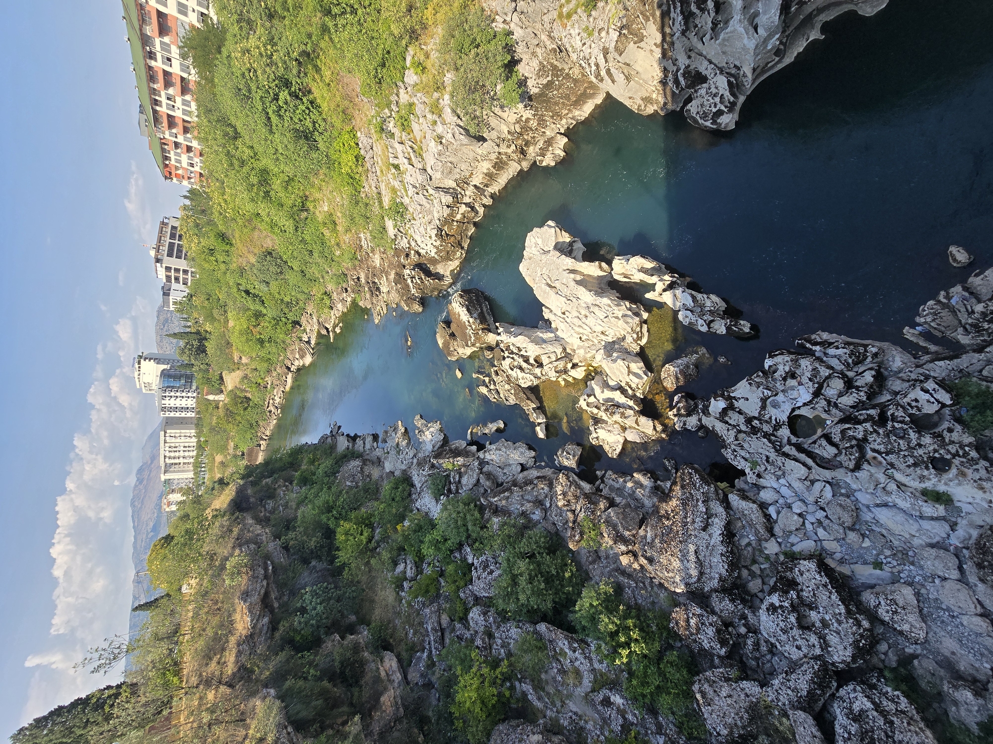 A high-angle, full shot captures a deep, narrow river cutting through a rocky gorge under a bright, clear sky with scattered white clouds. The river, a deep turquoise, flows around large, white, jagged rocks and boulders that line its banks and emerge from the water. The rocky terrain is covered in patches of green vegetation and shrubs. On the far bank, a steep, tree-covered hillside rises, leading to a cluster of modern buildings, including white, multi-story structures with balconies and a building with an orange and white facade and a green roof. In the distance, hazy mountains form the backdrop. The lighting suggests a sunny day, with strong shadows cast by the rocks and buildings. No people are visible in the scene.
