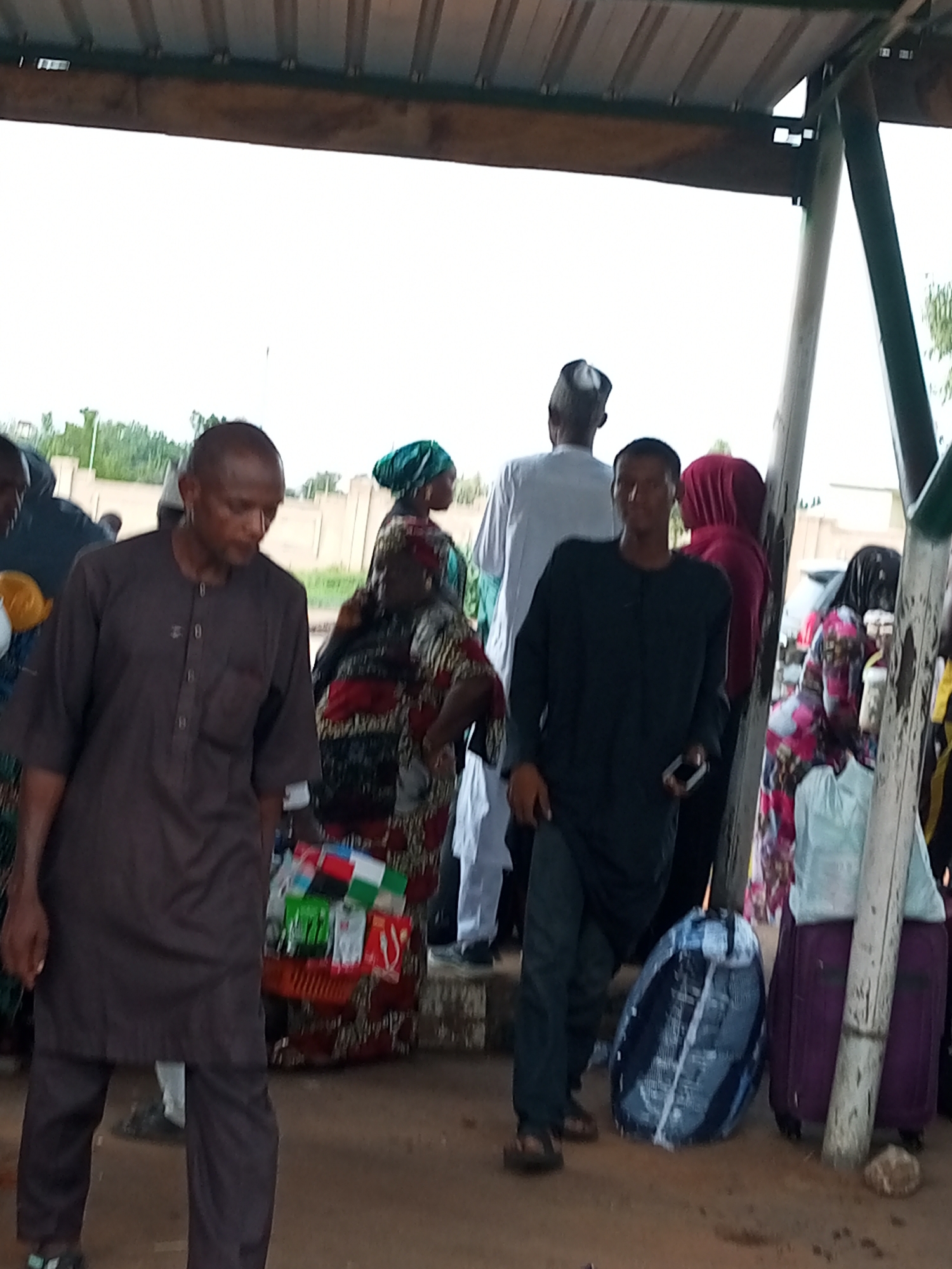 The image shows a group of people gathered under a shelter, likely a bus station or marketplace, in the city of Gombe, Nigeria. The shelter has a corrugated metal roof and wooden beams. In the foreground, a man wearing a dark brown traditional outfit is walking, looking down. To his right, another man in a black long-sleeved shirt and dark pants is also walking and holding a mobile phone. Behind them, several other individuals are visible, some in traditional attire. A stall with various items, possibly goods for sale, is positioned near the man in the brown outfit. Luggage, including a large round bag wrapped in plastic and a purple suitcase, are present. The ground appears to be dusty and uneven. The overall atmosphere suggests a busy transit point.