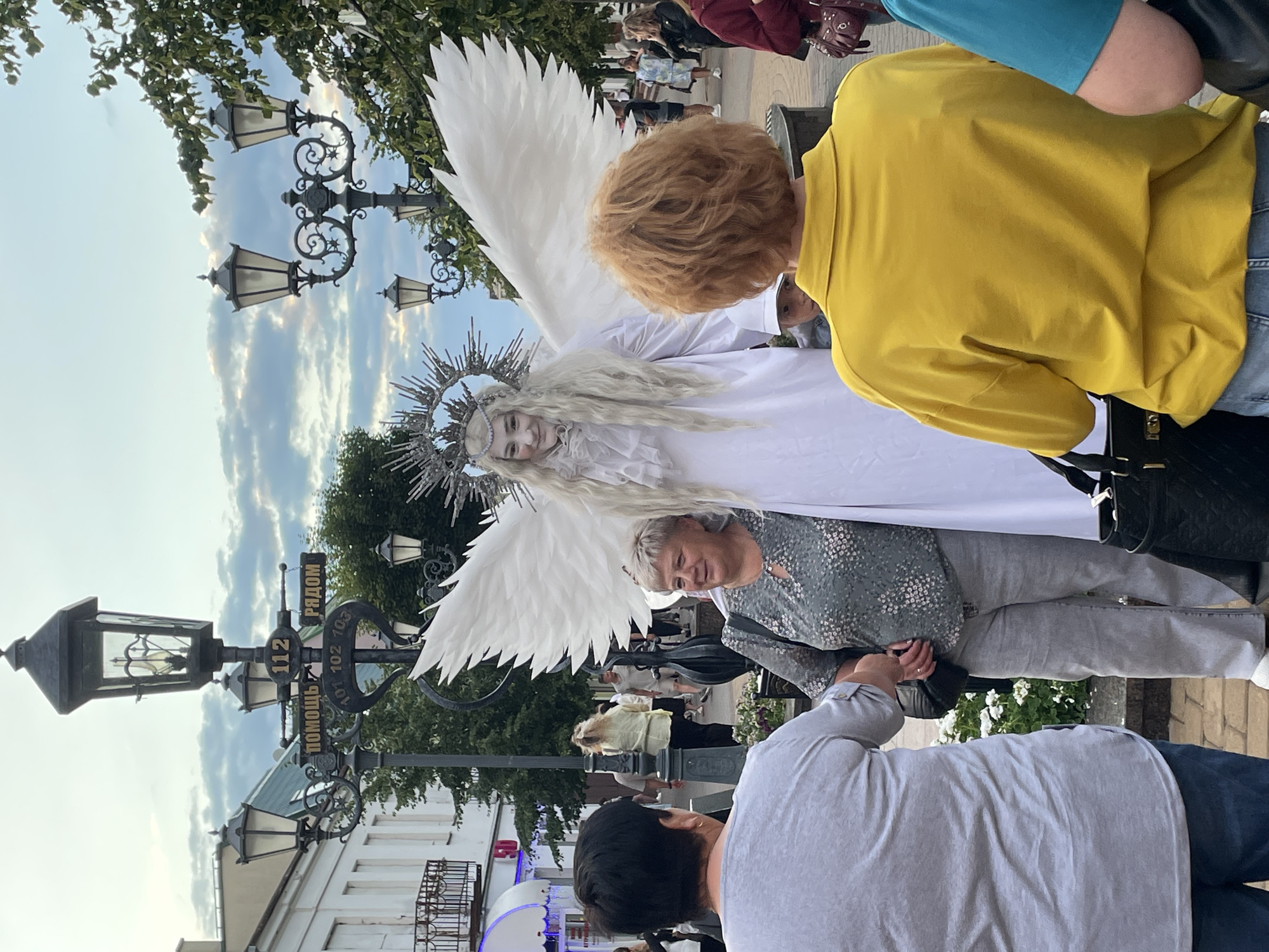 In the city of Brest, Belarus, on Sovetskaya Street, a street performer dressed as an angel with white feathered wings and a halo stands near a lamppost with street signs. Three women are interacting with the performer. One woman in a grey shirt and blue jeans is facing away from the camera, looking towards the performer. Another woman in a green patterned shirt and grey pants is smiling and looking at the performer. The third woman, seen from the back, wears a yellow shirt and blue jeans and is also looking at the performer. The environment includes buildings, trees, and other pedestrians in the background, under a cloudy sky. The lamppost displays signs with Cyrillic text, indicating street numbers and directions.