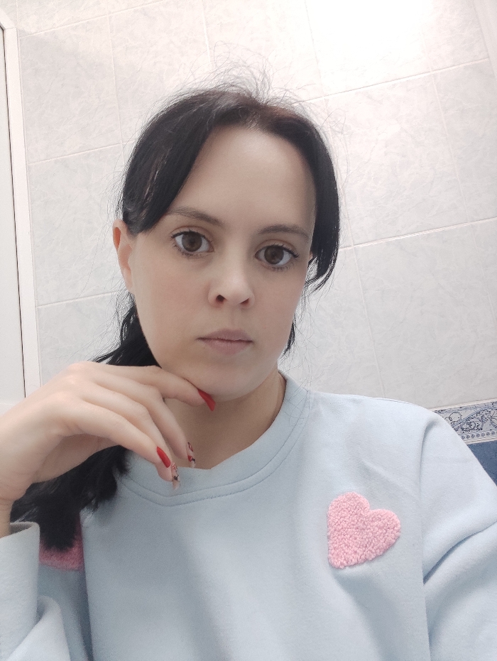 A female individual with dark hair, styled partially back, is depicted in a front-facing, close-up shot. Her brown eyes are directed towards the viewer, and she maintains a neutral expression. Her right hand, with red-painted fingernails, is raised with fingers lightly resting near her chin. She wears a light blue long-sleeved sweatshirt adorned with a textured pink heart-shaped patch on the left chest area. The background features light-colored, rectangular tiles with visible grout lines. A small, patterned blue and white object is partially visible in the upper right portion of the image. The specific location within the city of Donetsk, Ukraine, cannot be determined from the visual information provided.