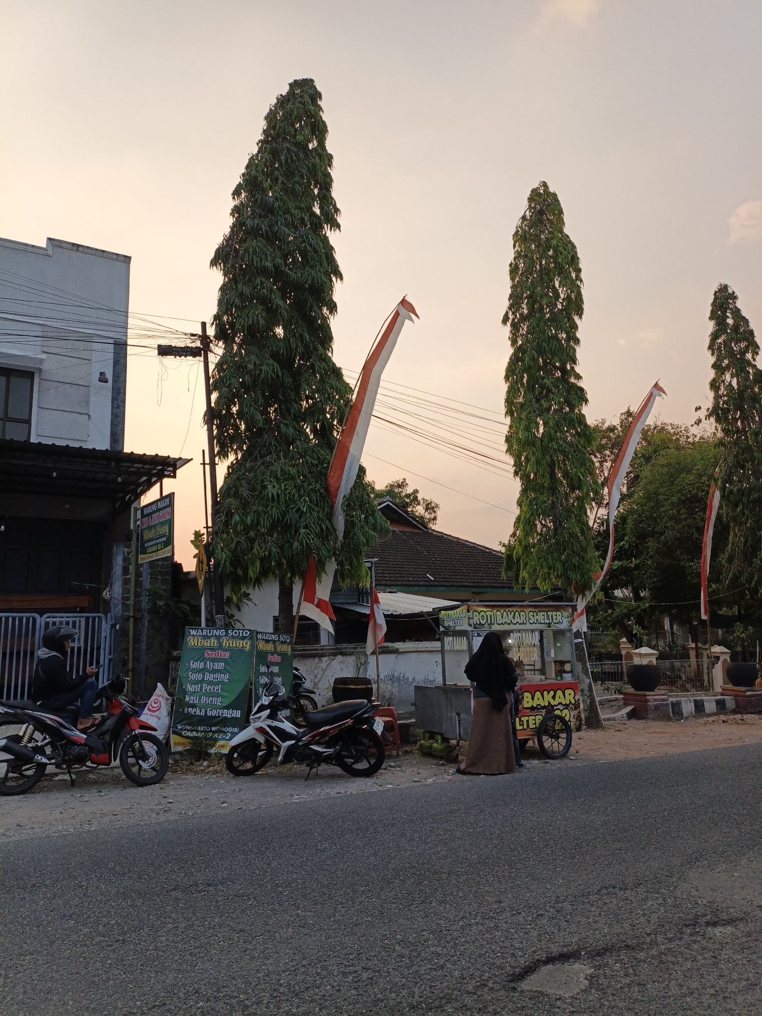 The image depicts a street scene at dusk in Wonokarto, Indonesia, with a focus on a food stall and its surroundings. Tall, slender trees with dense foliage dominate the background, interspersed with Indonesian flags. The sky is a pale orange, indicating the setting sun.
In the foreground, a paved road runs across the bottom of the image. To the left, a white building with a dark roofline and windows is visible. A covered area extends from this building, with metal bars in front. Two motorcycles are parked near this building. One is a red and black motorcycle with a rider wearing a helmet sitting on it, looking at their phone. Another is a white and black motorcycle parked next to it.
To the right of the building, a food stall with a red and yellow banner reading "ROTI BAKAR SHELTER" is situated. A woman wearing a black hijab and a long brown skirt stands behind the stall, facing away from the camera. In front of the stall, gas cylinders are visible. Next to the food stall, a sign in green reads "WARUNG SOTO Mbah Kung" followed by a list of food items including "Soto Ayam", "Soto Daging", "Nasi Pecel", and "Aneka Gorengan".
Further to the right, more trees and a white fence are visible. The overall scene suggests a typical street view of a residential or commercial area in Indonesia during the evening.