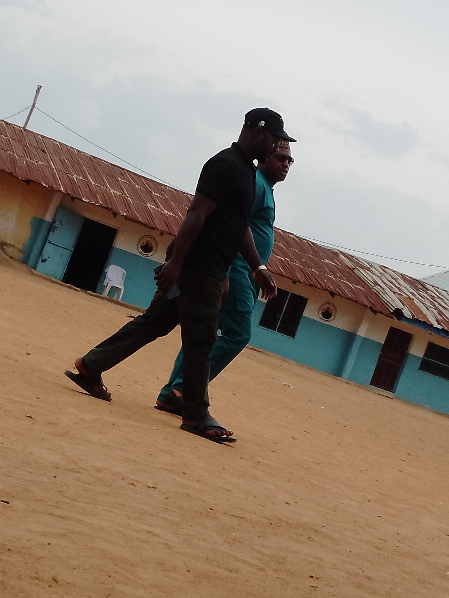 Two men are walking on a dirt path, likely a road, alongside a building. The man in the front is wearing a black t-shirt, dark pants, a black baseball cap, and sandals. He's holding a phone in his left hand. The man behind him is wearing a teal outfit, sunglasses, and a traditional hat. The building behind them is a light blue color with a rusted corrugated metal roof. The location is in the city of Unknown, Unknown.