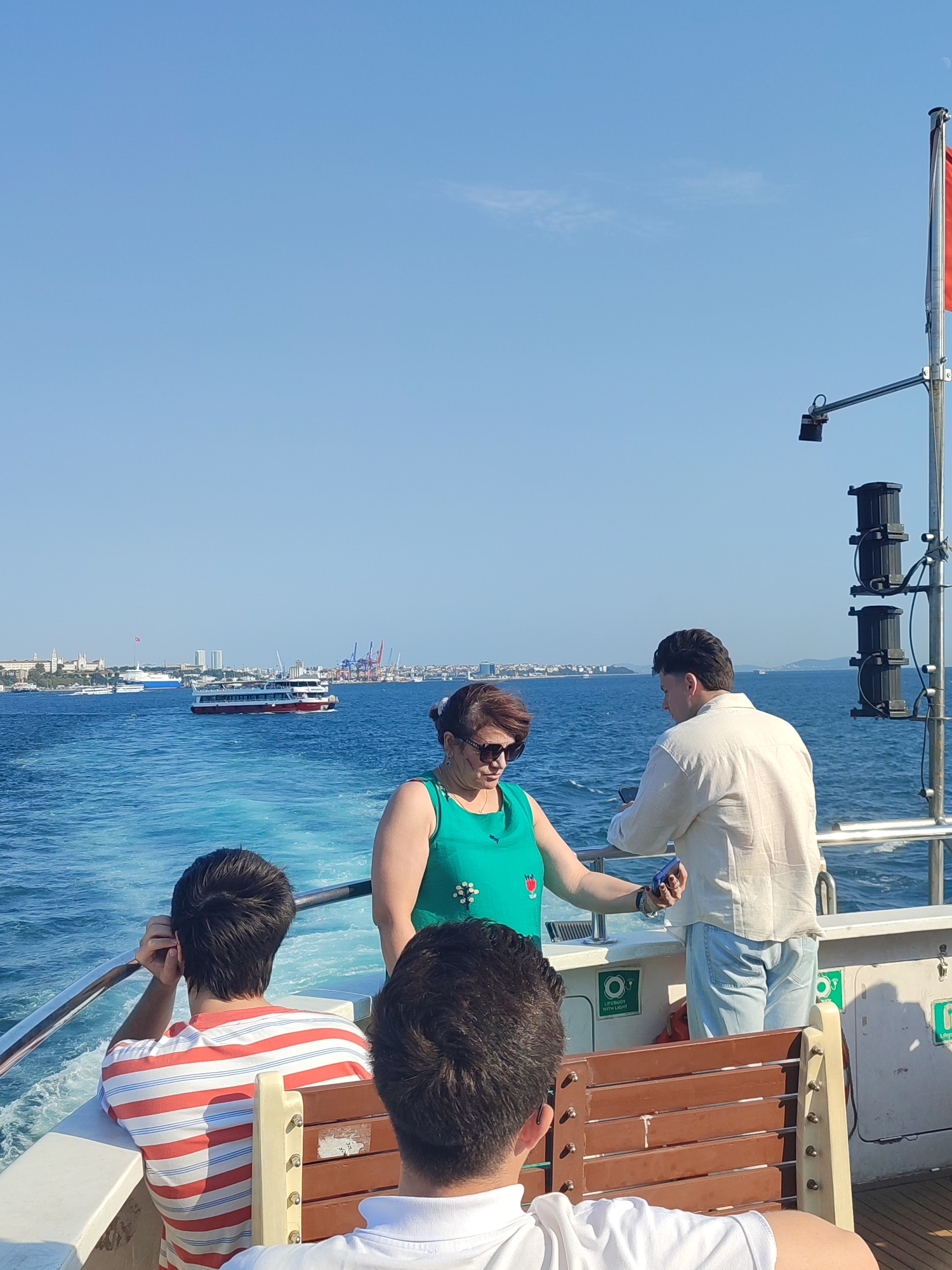 A medium shot captures a clear, sunny day on the Bosphorus Strait in Istanbul, Turkey. The foreground features the back of two men seated on a boat. The man on the left is wearing a red and white striped shirt, leaning against the boat's railing and looking out at the water. The man on the right is wearing a white polo shirt. In the mid-ground, a woman with dark hair and sunglasses, wearing a green dress with a floral pattern, is interacting with a man in a light-colored shirt and jeans. The man is looking at his phone. Behind them, the turquoise water of the strait churns with the wake of the boat. In the background, a passenger ferry with red and white detailing sails across the water. Further in the distance, the Istanbul skyline is visible, featuring buildings and industrial cranes, suggesting proximity to a port. A clear blue sky dominates the upper half of the image.