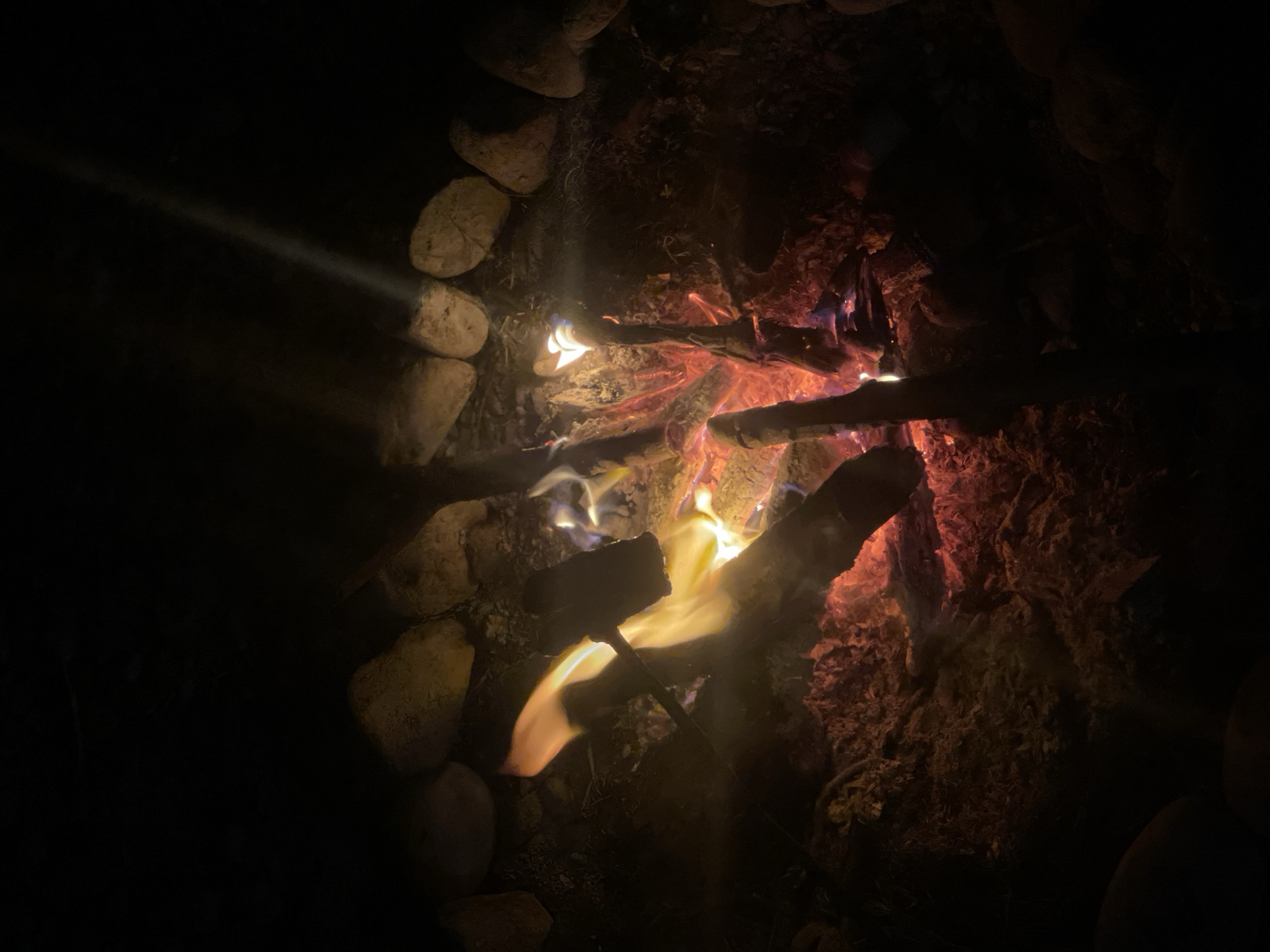 The image displays a close-up, low-angle view of a campfire at night. The fire is contained within a ring of light-colored stones. Several pieces of wood are burning, with flames ranging from yellow and orange to red, and hints of blue at the base. Embers and ash are visible on the ground beneath the fire. The surrounding area is dark, suggesting an outdoor setting. There is a narrow, bright beam of light descending from the upper right portion of the image, possibly from a light source outside the frame or a reflection. No people are present in the image. The location is specified as the city of Constance, Germany.