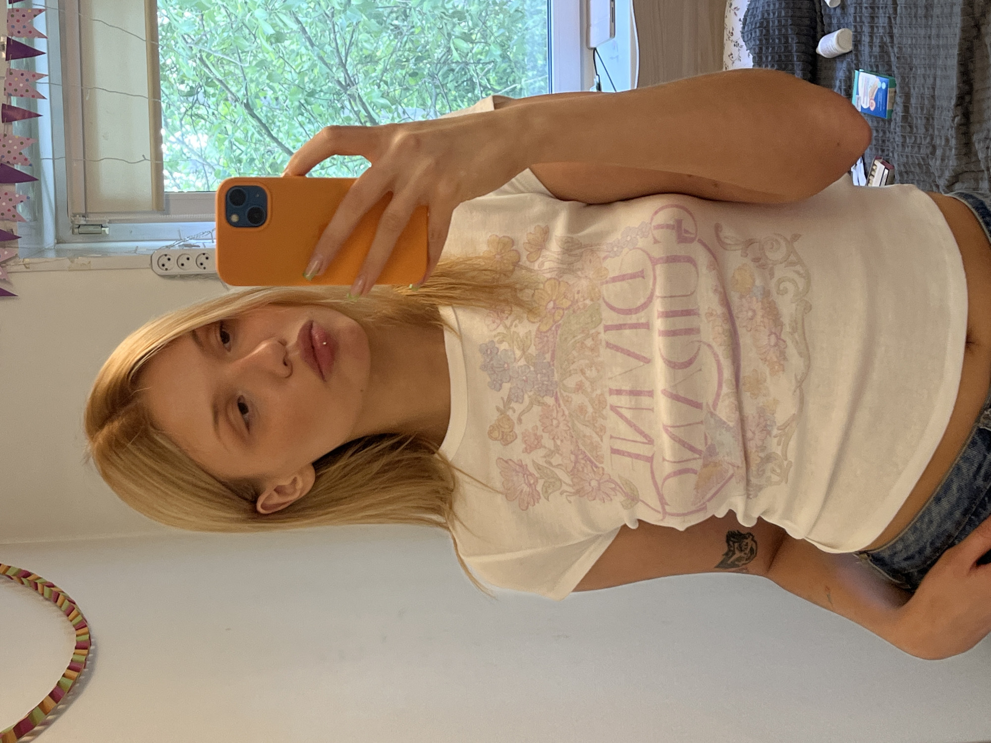 A young woman with blonde hair and a lip piercing is depicted taking a mirror selfie indoors, likely in Chaikovsky, Russia. She holds an orange smartphone with a triple-lens camera setup in her right hand, her nails adorned with light green tips. She is wearing a white short-sleeved t-shirt with a pastel floral design, featuring the word "DIVINE" in purple script across the chest. Part of a dark tattoo is visible on her right inner arm, just above the hem of her blue jeans.
The background reveals a brightly lit room, with a window on the left side looking out onto lush green trees, suggesting daytime. Below the window, a white power strip with three European-style outlets is mounted on the wall. To the far left, a decorative banner or garland with colorful triangular flags hangs vertically. On the right side of the image, behind the woman, is a gray textured surface, possibly a bed or blanket, with various small items scattered on it, including white bottles and a small blue box. A multi-colored circular object, possibly a hula hoop, is visible in the bottom left corner. The overall setting suggests a casual, personal space like a bedroom.