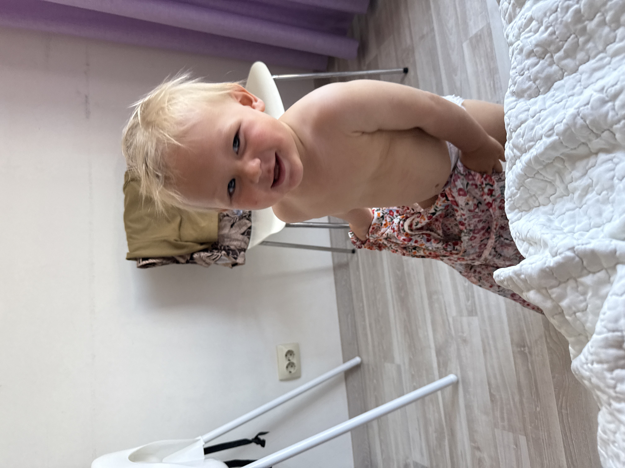The image shows a young child with blonde hair, appearing to be a toddler, seated on a white high chair. The child is wearing a white diaper and a colorful floral-patterned skirt or dress. The child is looking towards the camera with a slight smile and open mouth, seemingly engaged in an activity or posing.
Behind the child, a beige garment is draped over the high chair. The background consists of a white wall with a single electrical outlet. To the right of the child is a large white quilted blanket or bedspread. The flooring is light-colored wood laminate. In the upper part of the image, a purple curtain or fabric is visible.
The scene appears to be indoors, possibly a bedroom or a child's play area. The lighting suggests it is daytime. The child's expression conveys a sense of happiness or playful curiosity. The overall impression is a candid moment of a young child in a domestic setting. No visible text is present in the image.
