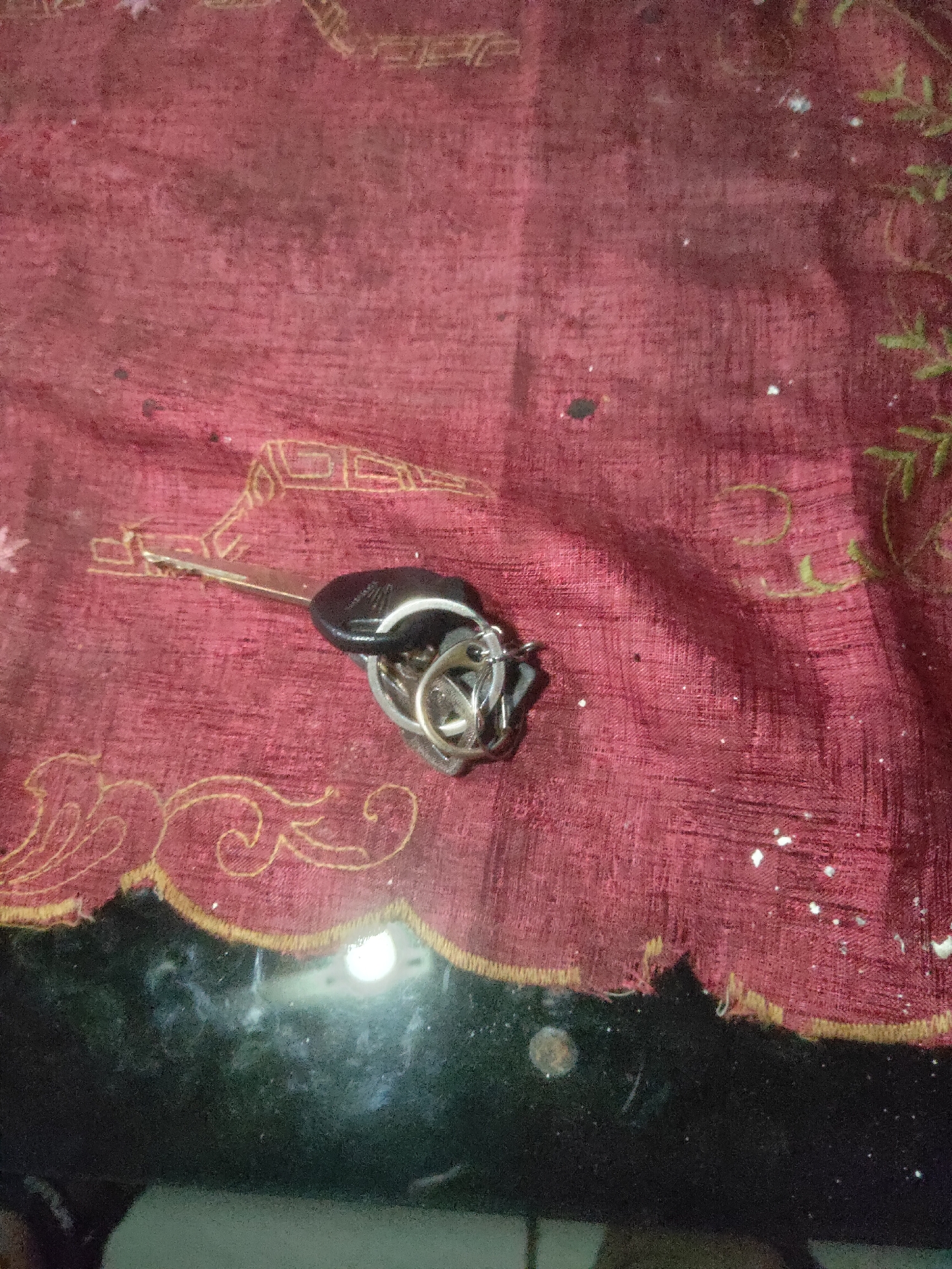 A bunch of keys with a black fob and a metallic keychain are lying on a red fabric surface with gold embroidery. The fabric has a woven texture and some scattered white specks. The keys are silver and appear to be in a slightly disordered pile on the fabric. The scene is dimly lit, with a bright, out-of-focus light source visible in the bottom portion of the image, suggesting a reflective surface like glass beneath the fabric. The location of Antirogo, Indonesia, is not discernible from the image itself.