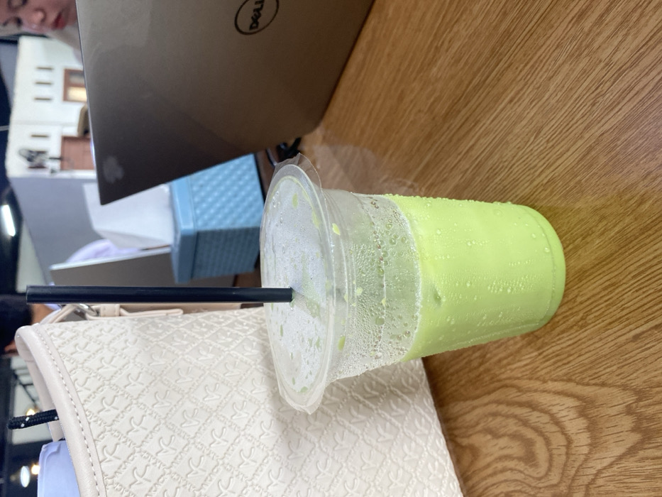 A clear plastic cup, partially filled with a light green, iced beverage and exhibiting condensation, stands on a light brown wooden table. A black straw is inserted through the plastic lid of the cup. To the left, a portion of an off-white, textured handbag with a patterned surface is visible. To the right, an open, bronze-colored Dell laptop is present on the table. Behind the laptop, a light blue, patterned tissue box is situated. A person's head and shoulder, wearing a light-colored headscarf, are partially visible in the upper right background, looking downwards. The scene takes place indoors, likely within a cafe or workspace, in Aluebutuha, Indonesia.