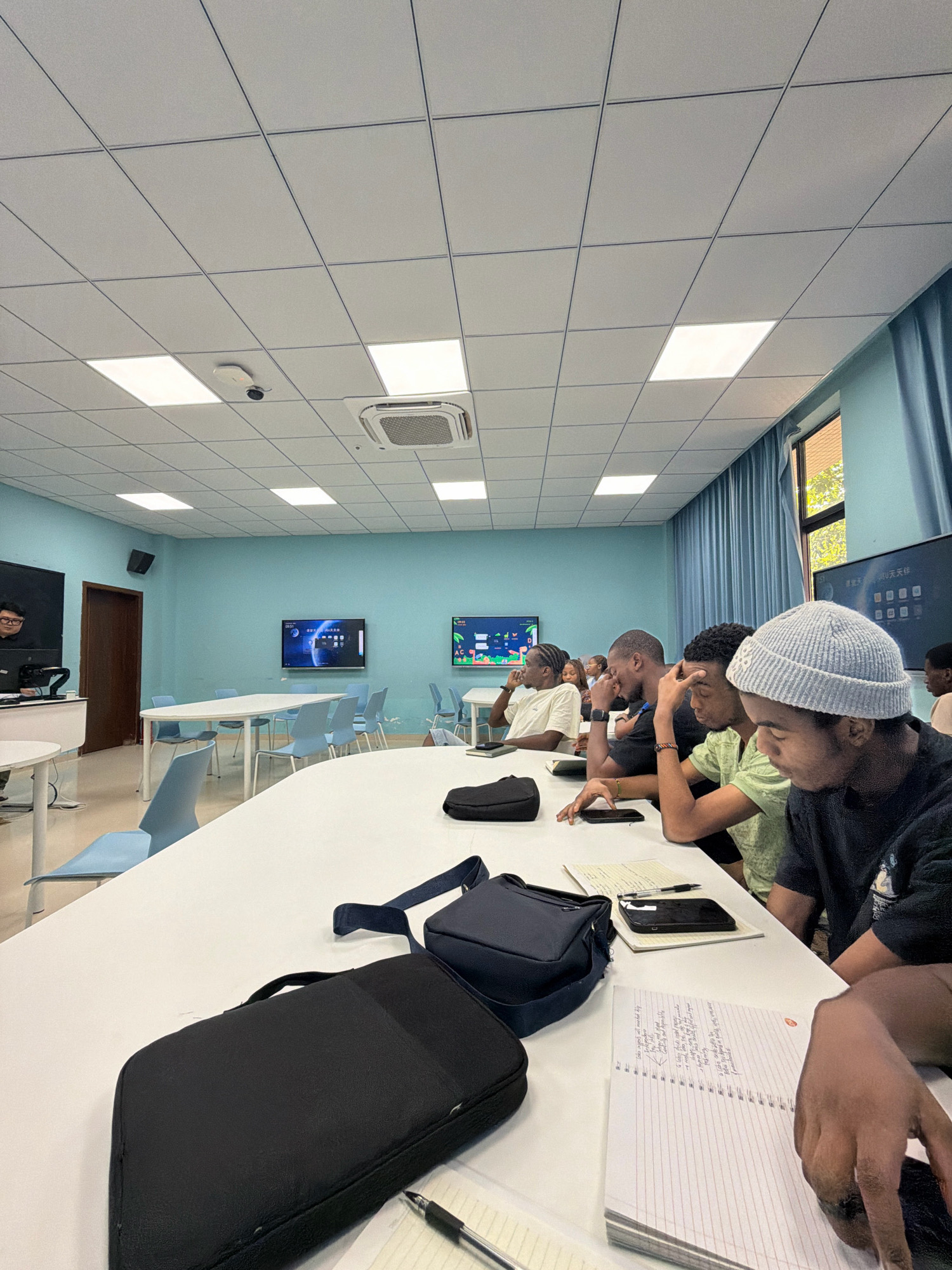The image depicts an indoor scene, likely a classroom or meeting room, within the city of Jiangjin, China. The room is brightly lit by overhead fluorescent lights and features a white, modular tiled ceiling with a visible air conditioning unit. The walls are painted a light blue, and a large window with blue curtains is present.
Several young individuals, appearing to be students, are seated at a long, white table. They are engaged in what appears to be a lesson or discussion. Some individuals are looking at their phones, while others are focused on notebooks and pens on the table. Several black bags are placed on the table and floor. In the background, large screens are mounted on the wall, displaying graphics and text. One individual, a man wearing glasses and a dark shirt, is visible standing at a podium with a laptop in front of a black board. The overall atmosphere suggests an educational or collaborative environment.