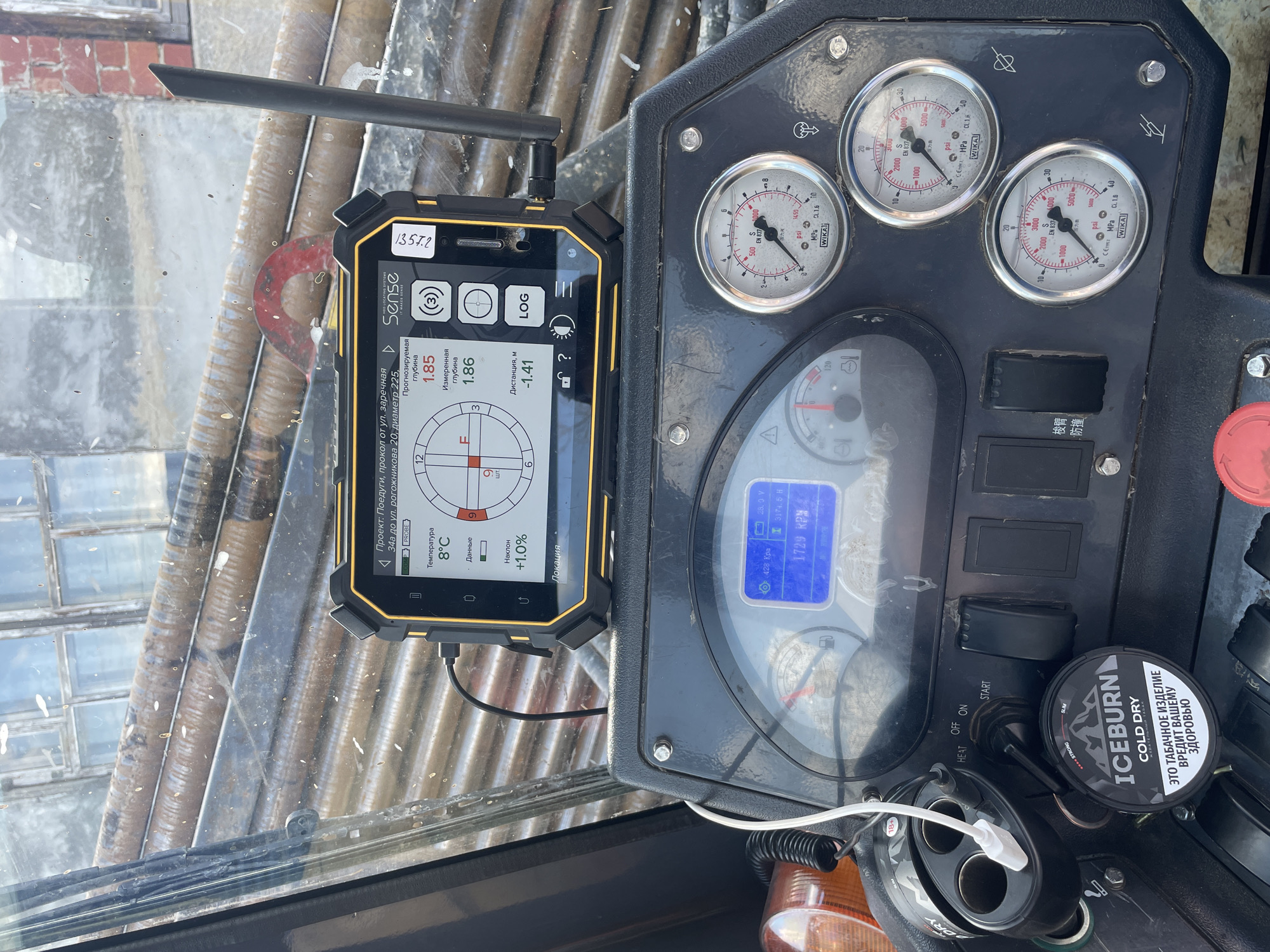 The image captures the dashboard of heavy machinery, possibly an excavator or drilling rig. Prominently displayed is a "Sense" brand tablet showing a drilling operation interface, indicating a planned depth of 1.85 meters and a measured depth of 1.86 meters. The screen shows the project location as "Поедуги, прокол от ул. заречная 34а до ул. рогожникова 20," suggesting the machine is operating at or near the intersection of Zarechnaya Street and Rogozhnikova Street in Поедуги, Russia. The tablet displays a compass-like interface with a +1.0% incline and a temperature of 8°C. To the right of the tablet, a cluster of analog gauges display pressure readings in psi and MPa, produced by "WIKA" brand. Below these, a digital display shows 1729 RPM. A tin of "ICEBURN Cold Dry" snus is visible near the vehicle's ignition. The surrounding area looks like the inside of a vehicle cabin. The overall image indicates ongoing drilling or excavation work at the identified location.