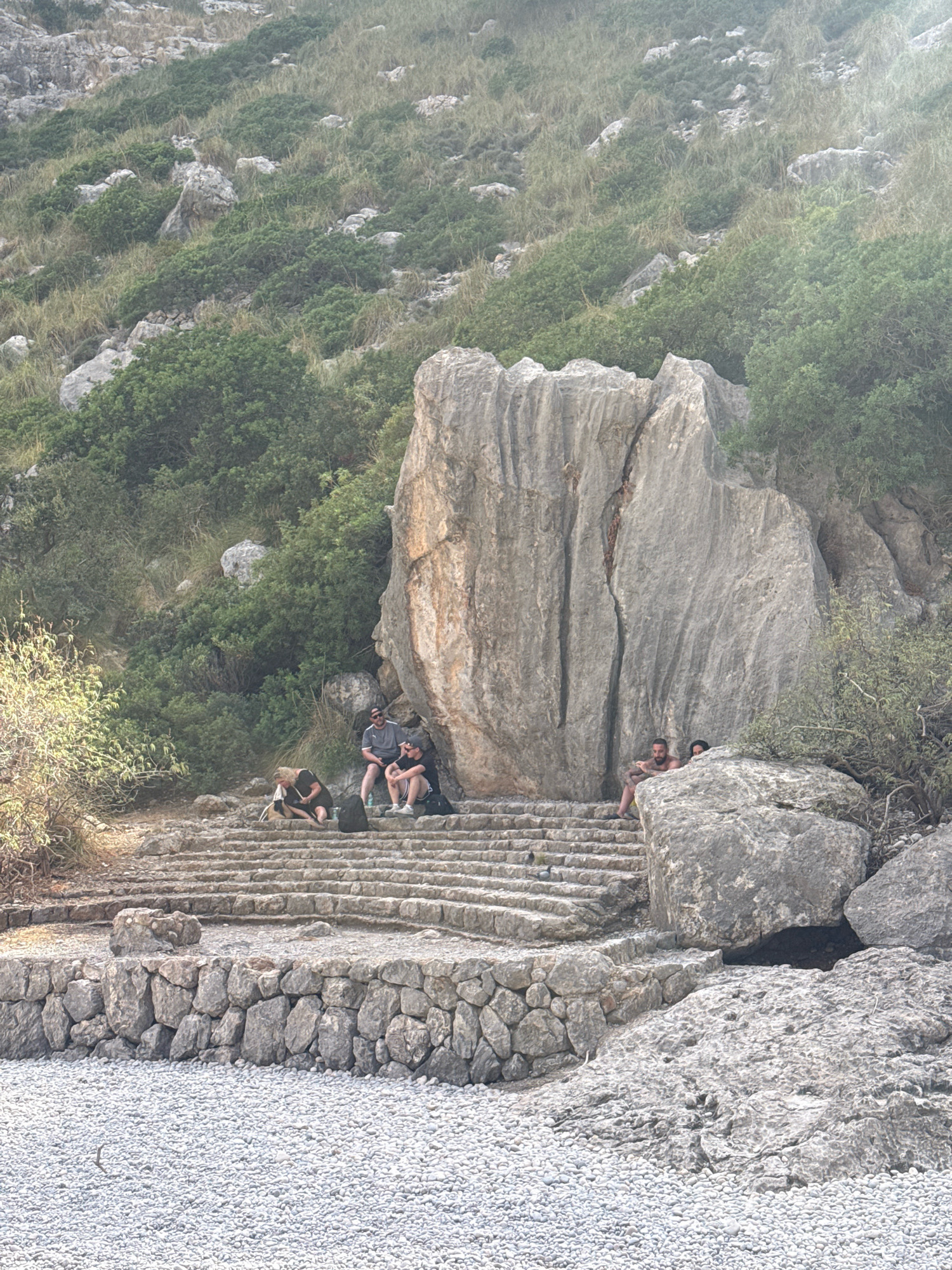 In Escorca, Spain, a group of people are resting on stone steps at the base of a rocky, vegetated mountainside. Several individuals are seated on the steps, some appearing to converse or relax. One person is seated on the ground near the steps. The environment consists of rough-hewn stone steps, a low stone wall, and a pebbled area in the foreground. A large, distinctive rock formation stands behind the steps, with lush green vegetation and rocky terrain filling the background.