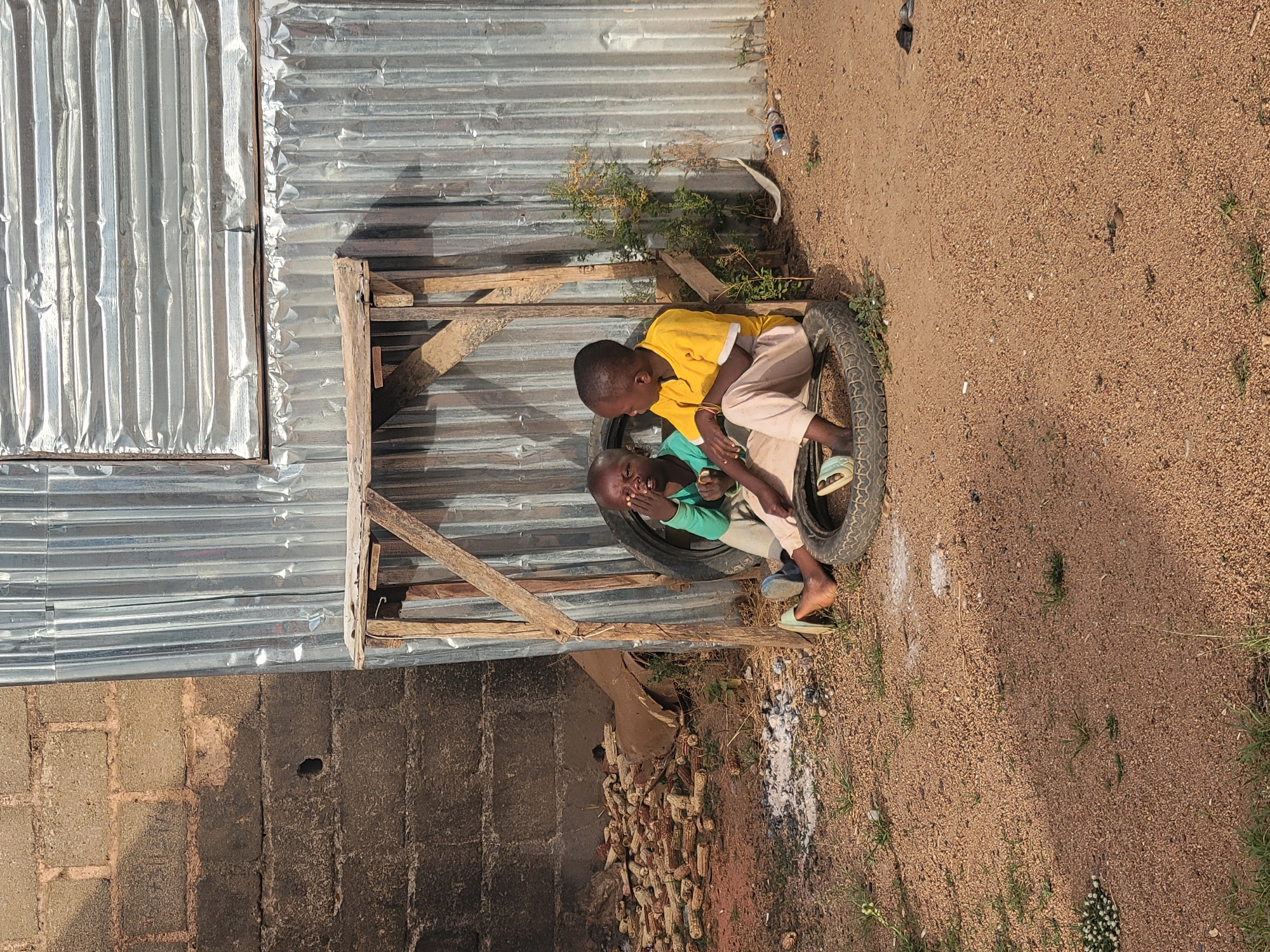 This image, taken in Barakin Lerawi, Nigeria, shows two young boys sitting on large rubber tires in an outdoor setting. The boy on the left is wearing a yellow short-sleeved shirt and light-colored trousers, looking down towards the other boy. The boy on the right is wearing a green short-sleeved shirt and similar light-colored trousers, with light blue sandals, and is looking towards the camera with an expressive, open-mouthed look, his hand near his face.
Behind the boys, a corrugated metal wall or shelter forms the backdrop, beneath which a dark block wall is partially visible on the left side. A simple wooden frame or stand is positioned in front of the corrugated metal. On the dry, sandy ground below the wooden frame and near the block wall, a collection of dried corn cobs is piled. The ground itself is composed of coarse dirt and small pebbles, with a few small green weeds growing sporadically. Further back on the ground, a discarded plastic bottle and other debris can be seen.
The scene is bathed in bright daylight, indicating a sunny and dry day. The shadows suggest it's either morning or late afternoon. The overall environment appears to be a humble, possibly rural or semi-urban area. The boys seem to be engaged in play or conversation, showing natural childhood interaction. No visible text is present in the image.
