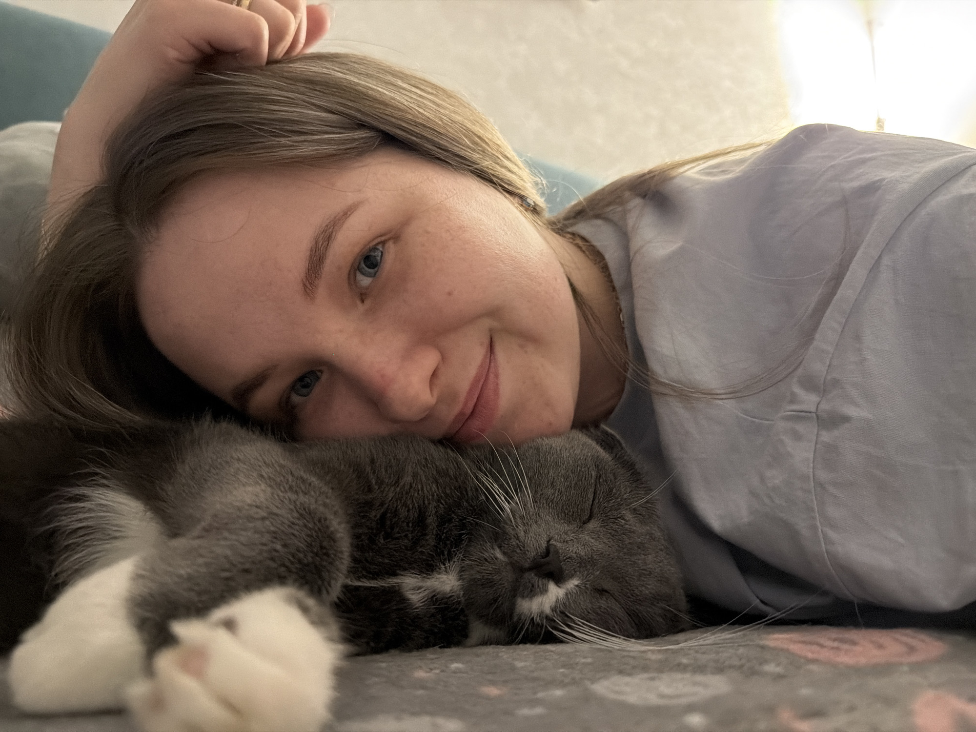 A woman is lying down with a gray and white cat. She is wearing a light blue top, and her head is resting next to the cat. The cat appears to be sleeping. They are lying on a gray blanket with a subtle pattern. The environment appears to be indoors, possibly a bedroom or living room. There is no information available in the image to indicate that this photo was taken in the city of Chaikovsky, Russia.