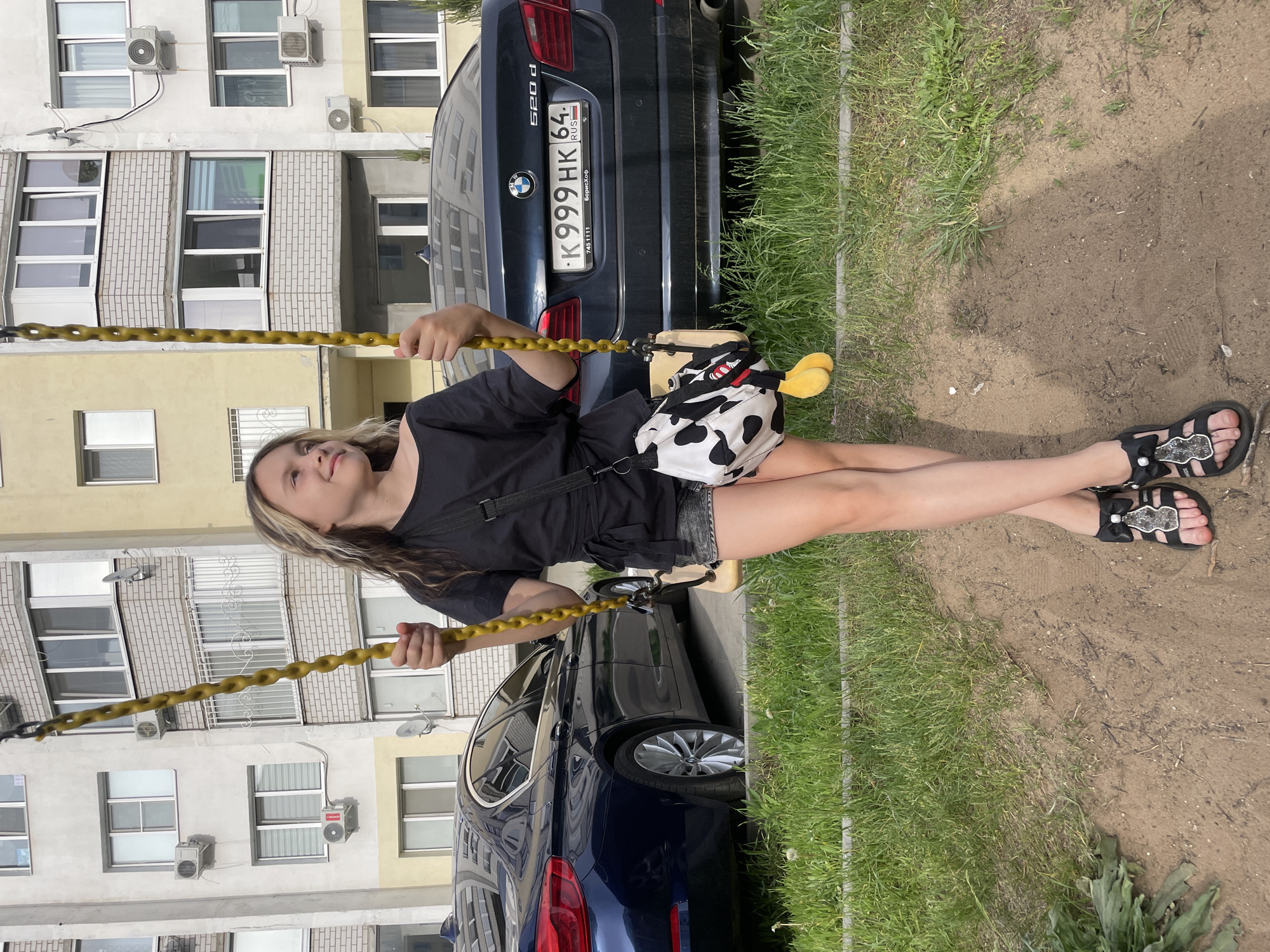 A young woman sits on a yellow swing next to a dark-blue BMW car. She is wearing a black t-shirt, grey shorts, and black sandals. The car has a license plate that reads "K 999 HK 64 RUS," which indicates it is registered in Russia. The swing is positioned on a patch of dirt and grass next to the car. In the background, there is a residential building with multiple windows and air conditioning units. The image seems to be taken in the city of Saratov, Russia.