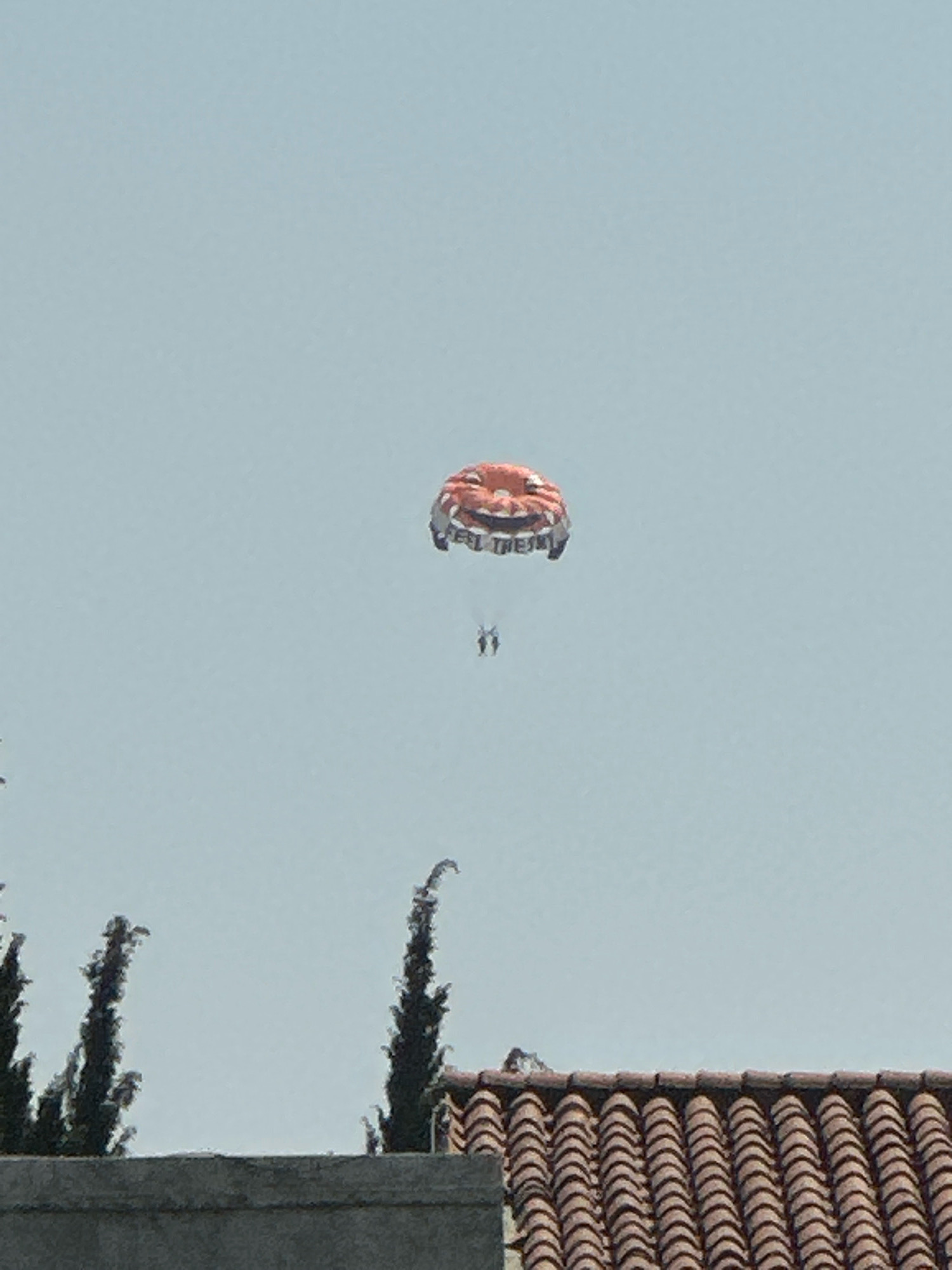The image shows a bright, overcast sky above a row of cypress trees and a tiled roof, suggesting a Mediterranean or coastal environment. In the upper center of the frame, a parasailer is visible, suspended by a large, orange and white parachute adorned with the words "FEEL THE SKY". Two people are clearly visible under the parachute, appearing to be enjoying the activity. The foreground features the edge of a concrete structure and the top of a red-tiled roof, implying a residential or urban area. The cypress trees, common in coastal regions of Montenegro, and the architectural style of the roof suggest the location could be Budva, Montenegro.