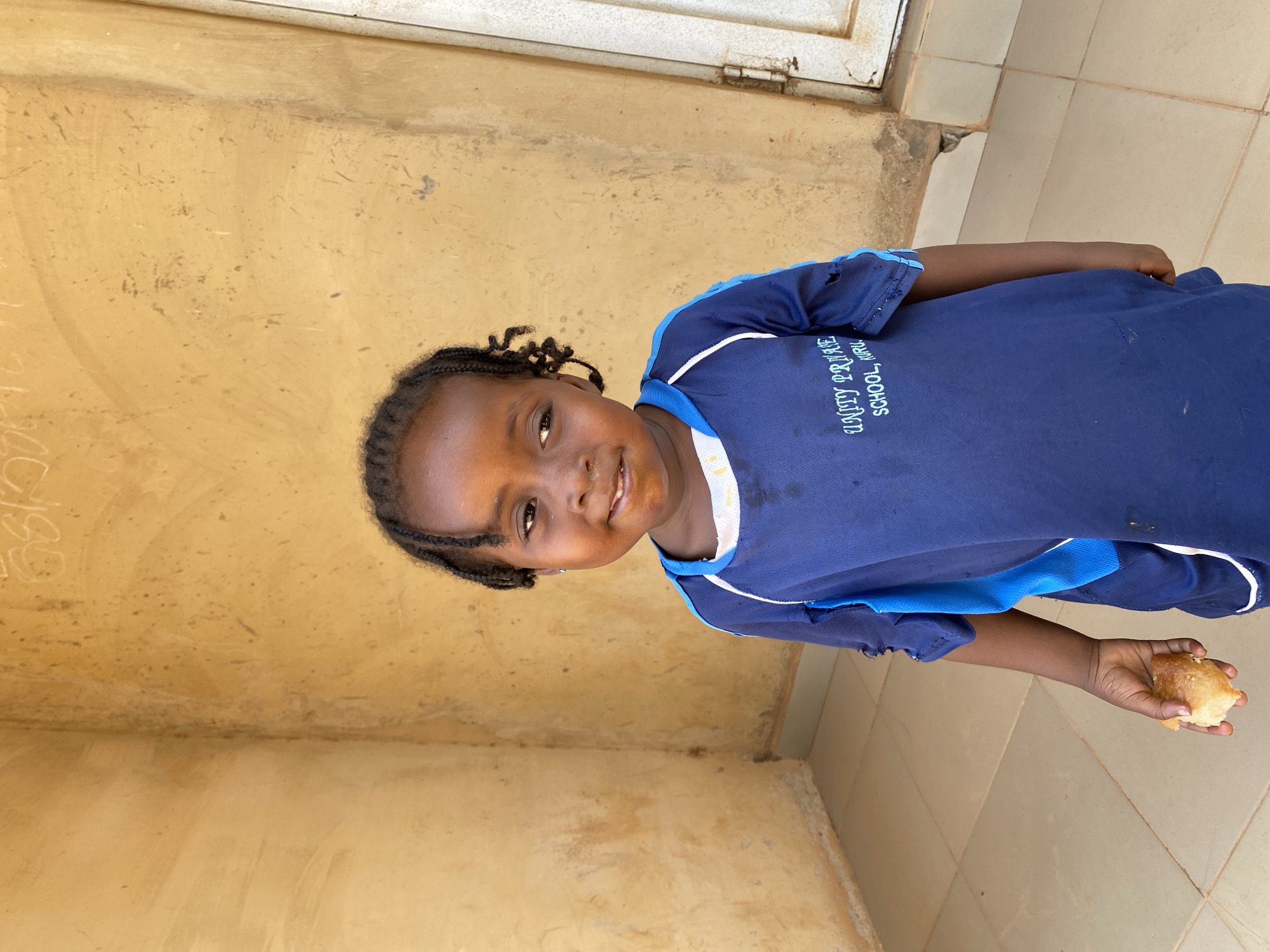 A young girl stands near a textured, yellow wall and tiled floor in Kuru Dungus, Nigeria. She wears a blue school uniform with "UNITY PRIVATE SCHOOL, KURU" printed on the front and holds a piece of bread in her right hand.