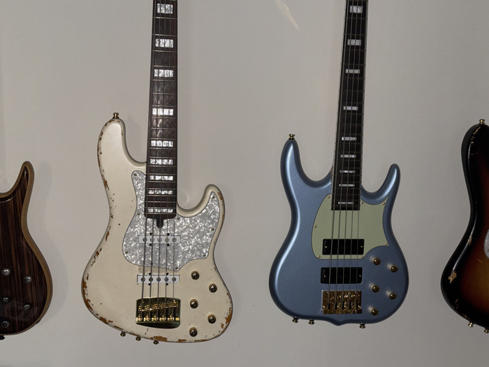 Four bass guitars are mounted vertically against a plain, light-colored wall in what appears to be an indoor setting. From left to right:
1. A partial view of a bass guitar's body in a dark wood finish, likely zebrawood or similar, displaying prominent grain.
2. A four-string bass guitar with a heavily distressed, off-white or cream finish, showing wear and tear revealing dark wood underneath. It features a white pearloid pickguard, two J-style pickups, a gold-colored bridge, and six gold control knobs. The fretboard has rectangular block inlays.
3. A five-string bass guitar with a metallic light blue finish. It has a light green pickguard, two black pickups (likely P/J or humbucker configuration), a gold-colored bridge, and six gold control knobs. The fretboard also features rectangular block inlays.
4. A partial view of a four-string bass guitar's body with a sunburst finish (dark brown/black fading into red/orange), also showing signs of significant wear.
No individuals are present. The guitars are static, mounted for display or storage. The specific location within the city of Los Angeles, United States of America, cannot be determined from the visual content of the image alone; however, the scene is consistent with a music store, a private collection, or a recording studio.