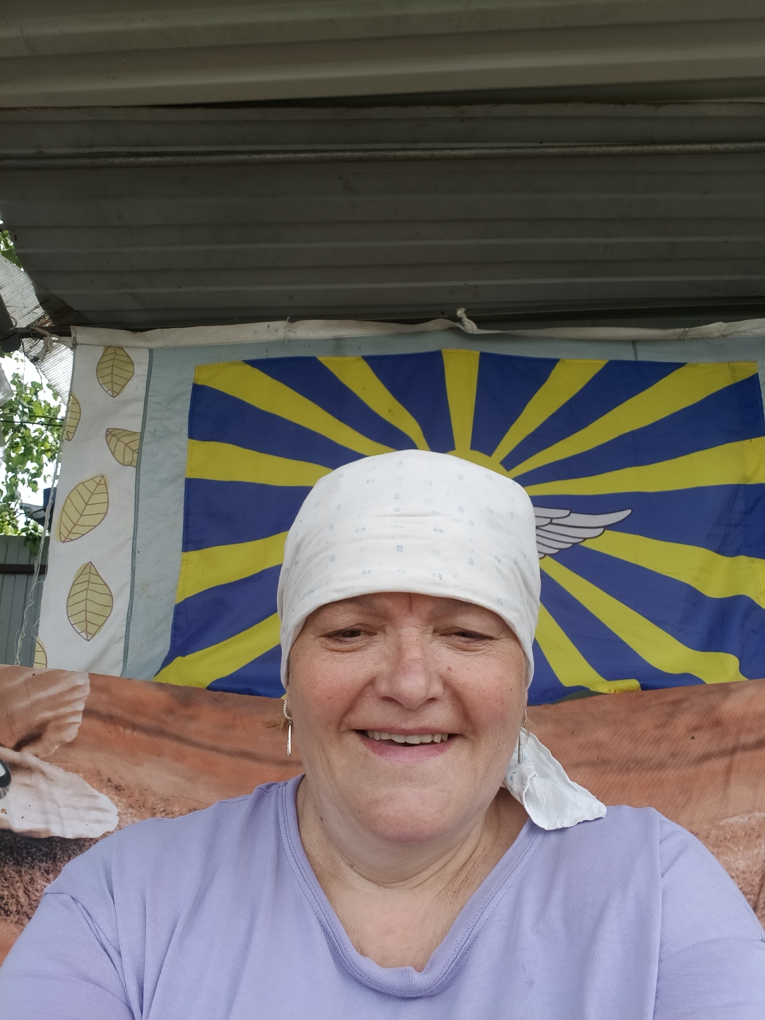 A woman wearing a white headscarf and a light purple t-shirt smiles at the camera. She has fair skin and short hair peeking out from under the headscarf. Behind her is a backdrop of various fabrics and materials, including a blue and yellow striped flag-like banner with a white wing emblem. There's also a cloth or banner with a brown, possibly animal-themed pattern, and another white banner with leaf-like designs. The setting appears to be outdoors under some sort of corrugated metal roofing, possibly a makeshift shelter. This image was taken in Красный Маяк, Russia.