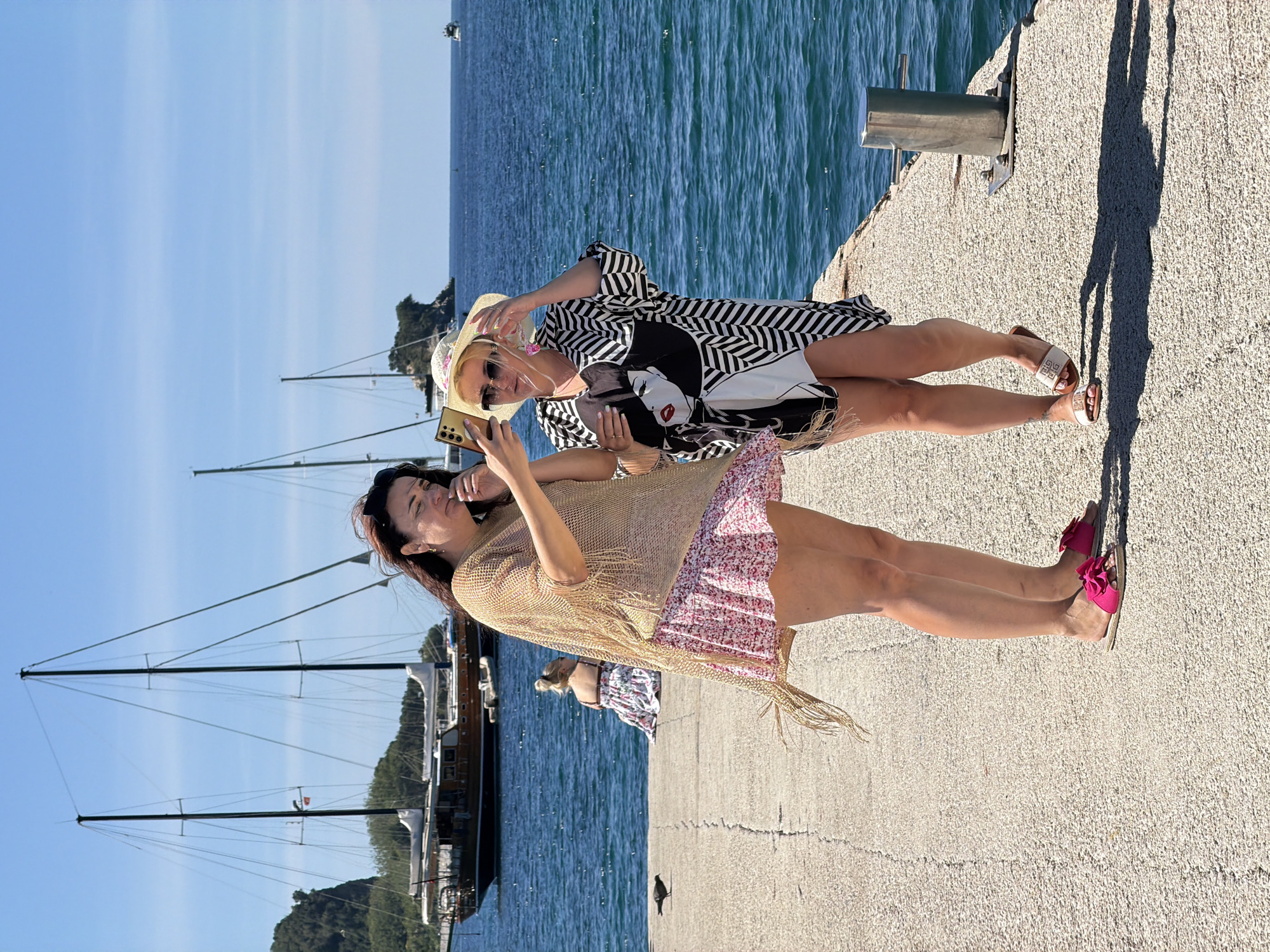 Two women are taking a selfie on a concrete pier against a backdrop of the Adriatic Sea, boats, and distant land. The woman on the left has dark hair, a light tan shawl, and a floral print dress. She holds a gold phone to take the photo. The woman on the right wears a straw hat, sunglasses, and a black and white geometric print dress. She holds up two fingers in a peace sign. The pier has a metal post. Their shadows stretch out on the pier. The location is likely the city of Budva, Montenegro, given the typical coastal landscape and architecture.