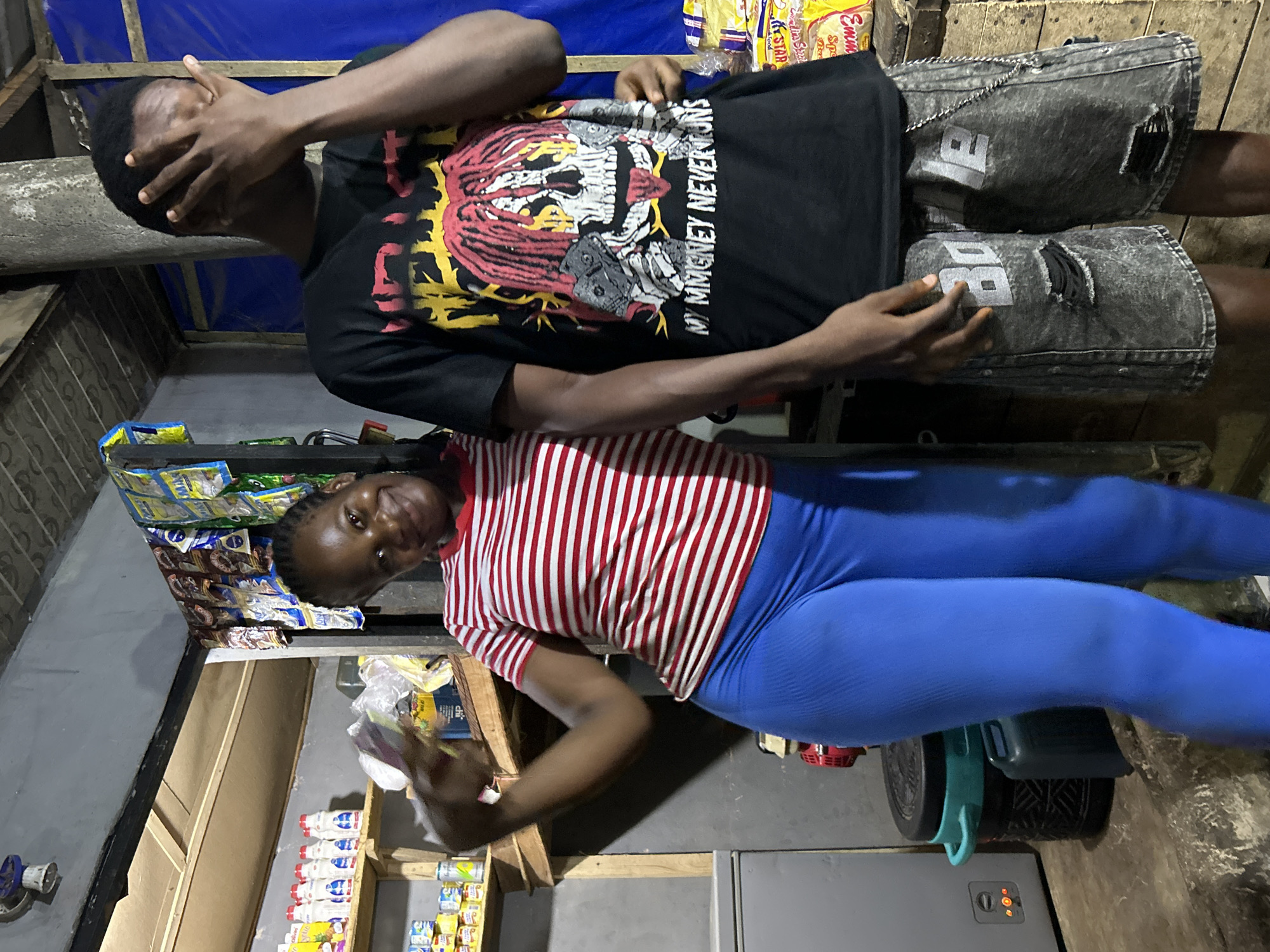 Two individuals are present in the image, possibly within or adjacent to a small convenience store in Ihiagwa, Nigeria. The store shelves are stocked with various food items and household products. One person stands partially behind the other, obscuring their face with their hand. The individual behind is wearing a black t-shirt with a graphic print and gray denim shorts with visible distressing. The person in front is wearing a red and white striped shirt and blue leggings. The setting appears modest, suggesting a local business within the Ihiagwa community.