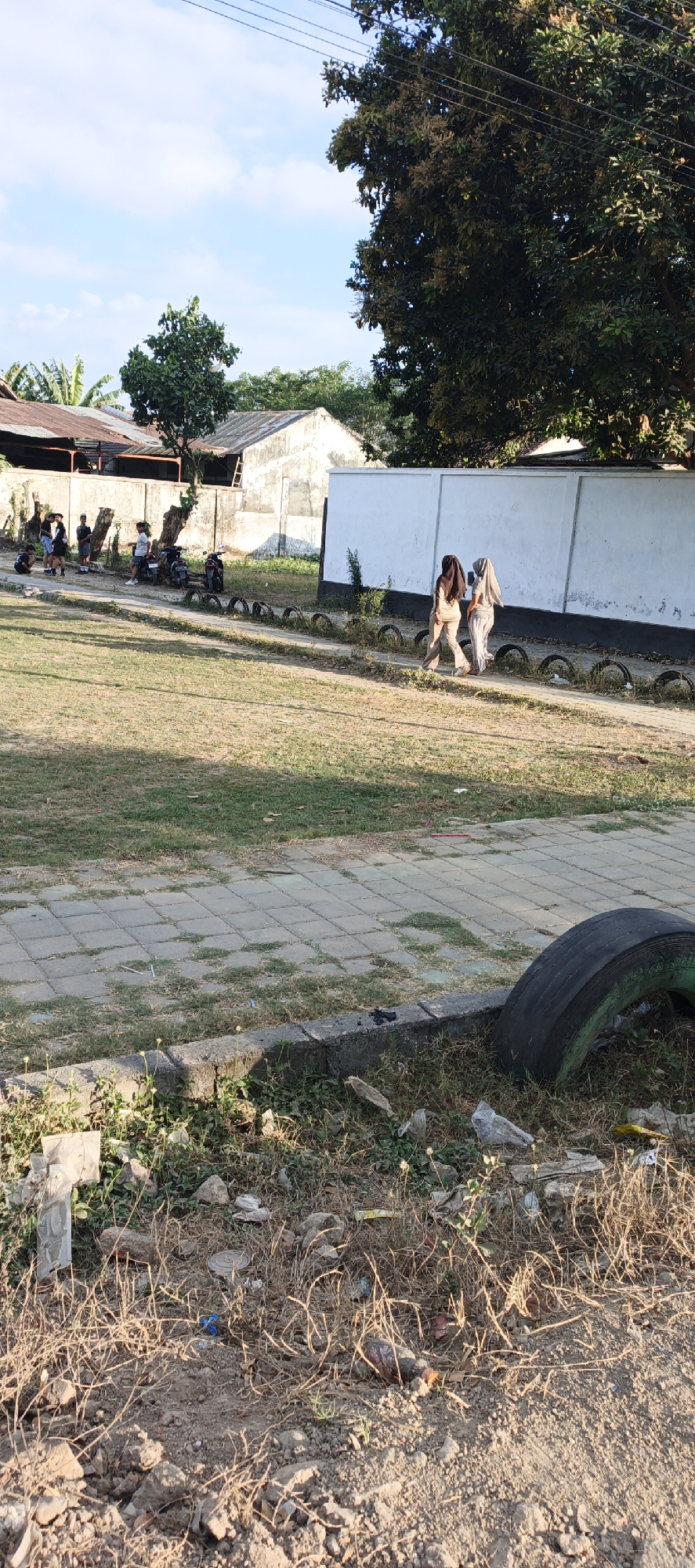 In Kasiyan Timur, Indonesia, a sunny daytime scene unfolds in an outdoor area with a paved walkway and grassy areas. Two individuals, wearing hijabs and long clothing in muted colors, are walking on the walkway. In the background, several motorcycles are parked near a white wall. Adjacent to the walkway, a line of car tires is visible. The ground is covered with grass and some debris. The sky is bright blue with scattered clouds.