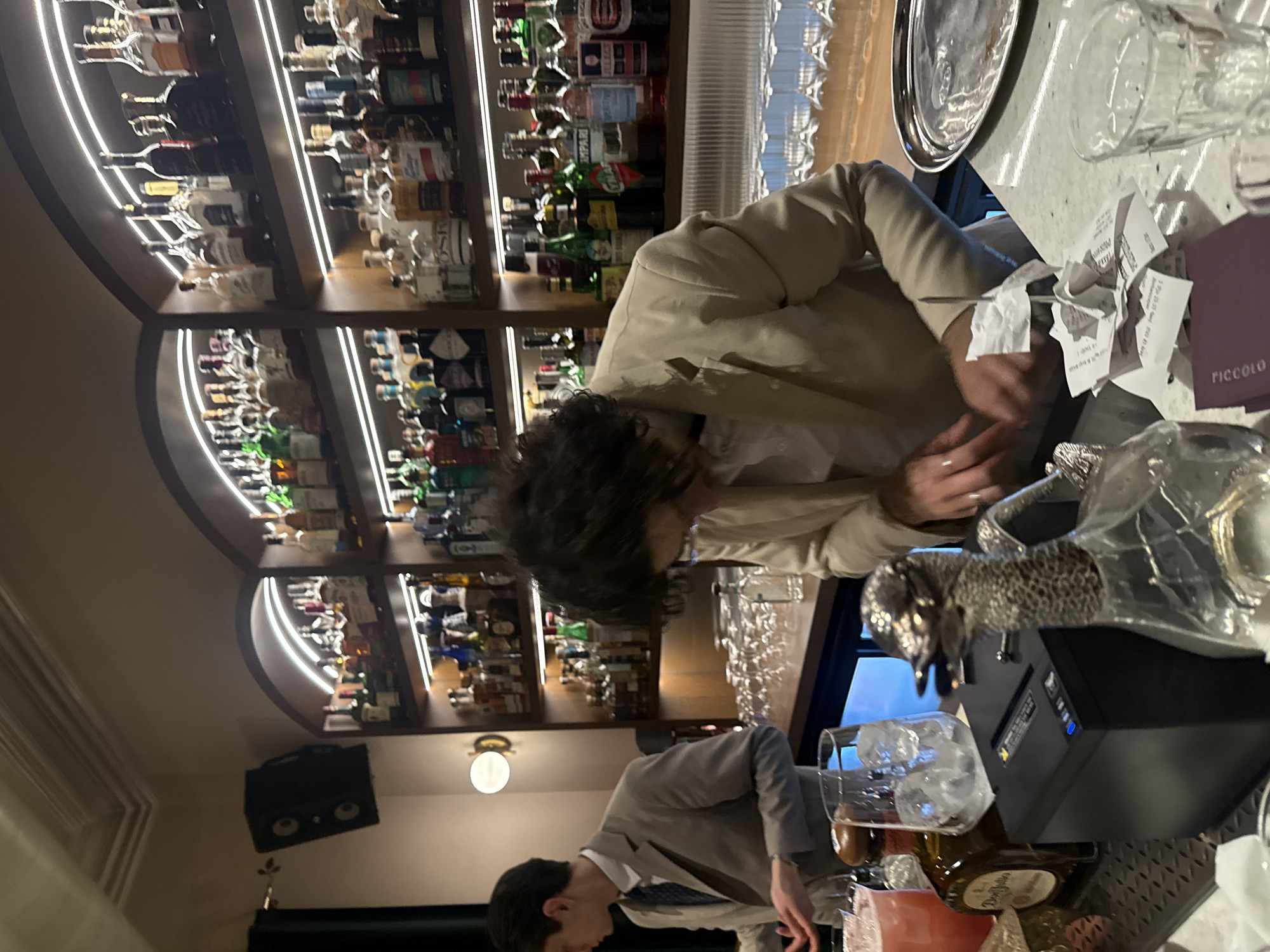 The image captures a bar scene with two individuals in Yerevan, Armenia. Behind them, a lighted liquor cabinet showcases various bottles. One individual, wearing a light-colored jacket, appears to be interacting with receipts on the bar top. Next to them, the edge of a purple 'Piccolo' menu is visible. In the foreground, a man in a gray suit is visible with a beer or cocktail and a black dispenser, possibly for drinks or ice, in front of him. The environment appears to be a well-lit bar or restaurant.