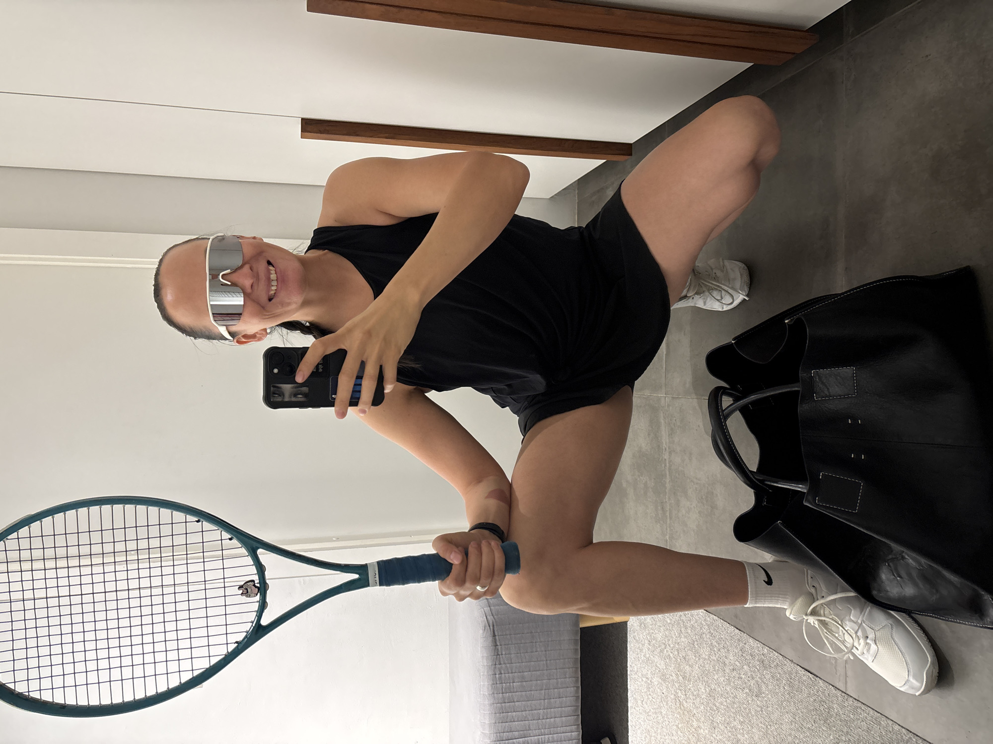 A woman is taking a selfie in a mirrored room. She is wearing silver reflective sunglasses, a black tank top, and black shorts. She is holding a tennis racket in her left hand and a cell phone in her right. The woman is squatting, with one leg forward and the other back. A black tote bag and white sneakers are near her feet. The room appears to have a gray tile floor, white and wood paneling on the wall, and a bed in the lower portion of the frame. There is no specific indication that the photo was taken in Chaikovsky, Russia.