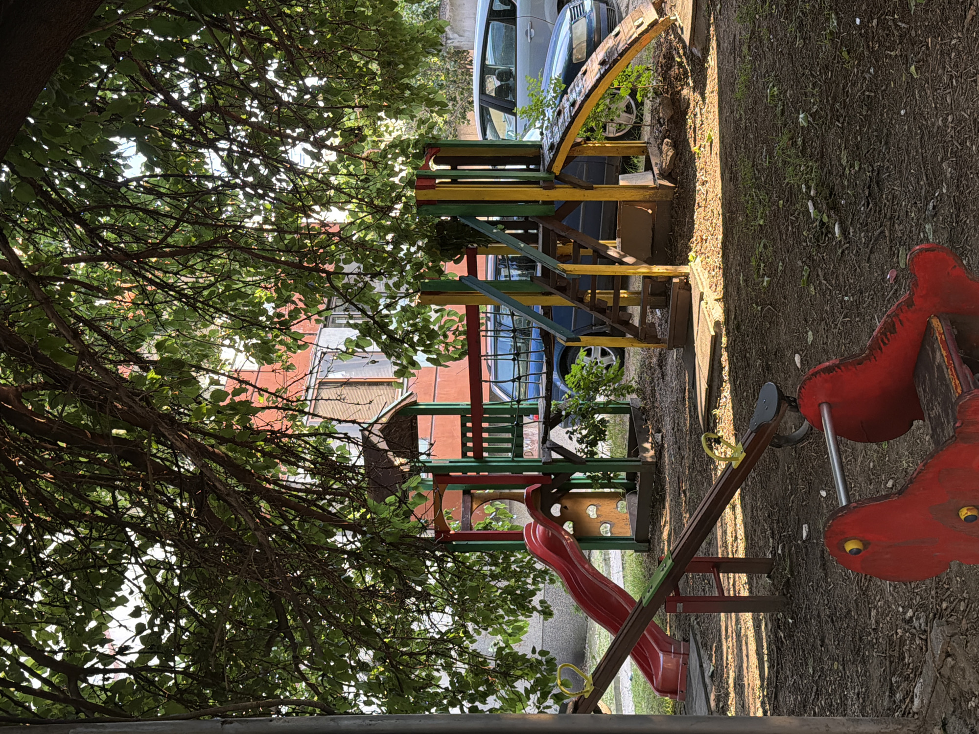 The image depicts an outdoor children's playground situated in Pazardzhik, Bulgaria. The playground features a red slide, a seesaw, and a multi-colored play structure with yellow stairs and a wooden climbing ramp. The play equipment is set against a backdrop of a building with reddish-orange walls and windows, partially obscured by the dense green foliage of a large tree overhead. Several vehicles, including a silver station wagon and a darker colored car, are parked in the vicinity. The ground is covered with dirt, scattered leaves, and some sparse vegetation. The scene is illuminated by sunlight filtering through the tree branches, casting dappled shadows. There are no people visible in the image.