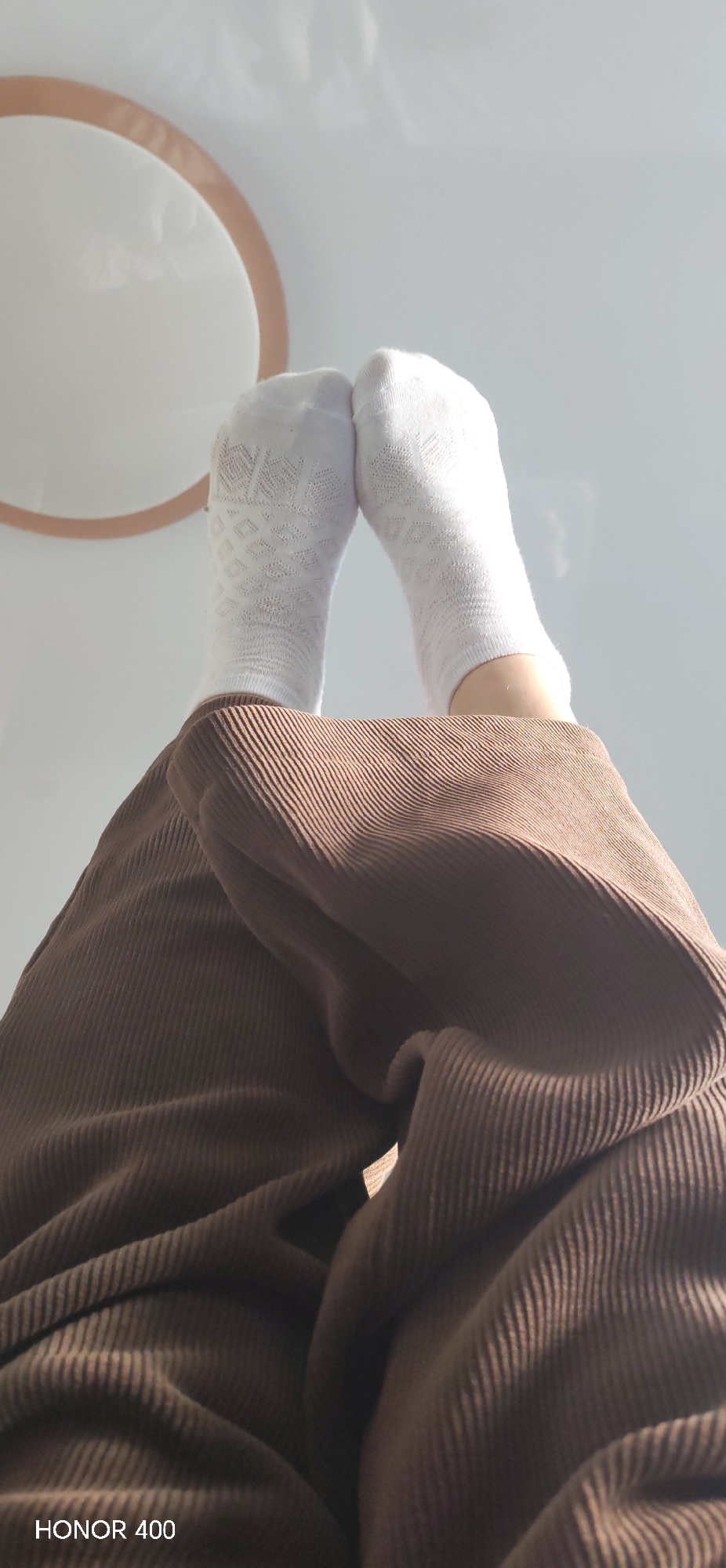The image shows a view from the perspective of someone lying down, looking up at their feet. The person is wearing brown corduroy pants and white socks. One sock has a patterned design. Above the feet, a round object with a light tan frame and white center is visible. The background is a light color, possibly a ceiling or wall. The text "HONOR 400" is present in the lower left corner of the image. The location (Noviy, Russia) cannot be determined from the visual information in the image.