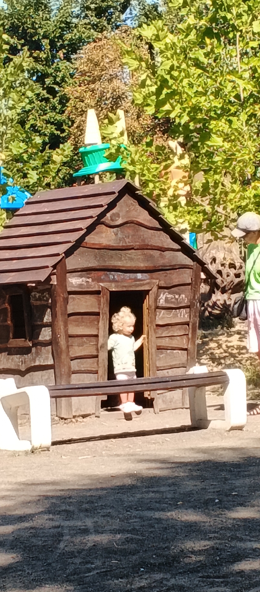The image captures a child with blonde, curly hair standing in the doorway of a rustic wooden playhouse. The child is wearing a light-colored t-shirt and pink shorts. To the left of the playhouse, a white bench with a dark brown top is partially visible. In the background, lush green trees and foliage form a dense canopy. To the right, an adult wearing a light-colored cap and a green shirt is visible, partially obscured by the trees. A textured, dark object resembling a rock or tree stump is also present to the right of the playhouse. The ground in front of the playhouse appears to be a mixture of dirt and gravel. The scene is set outdoors on a sunny day, suggesting it is likely a park or playground. There is no visual information within the image that allows for the determination of its specific location within the city of Cherkasy, Ukraine.