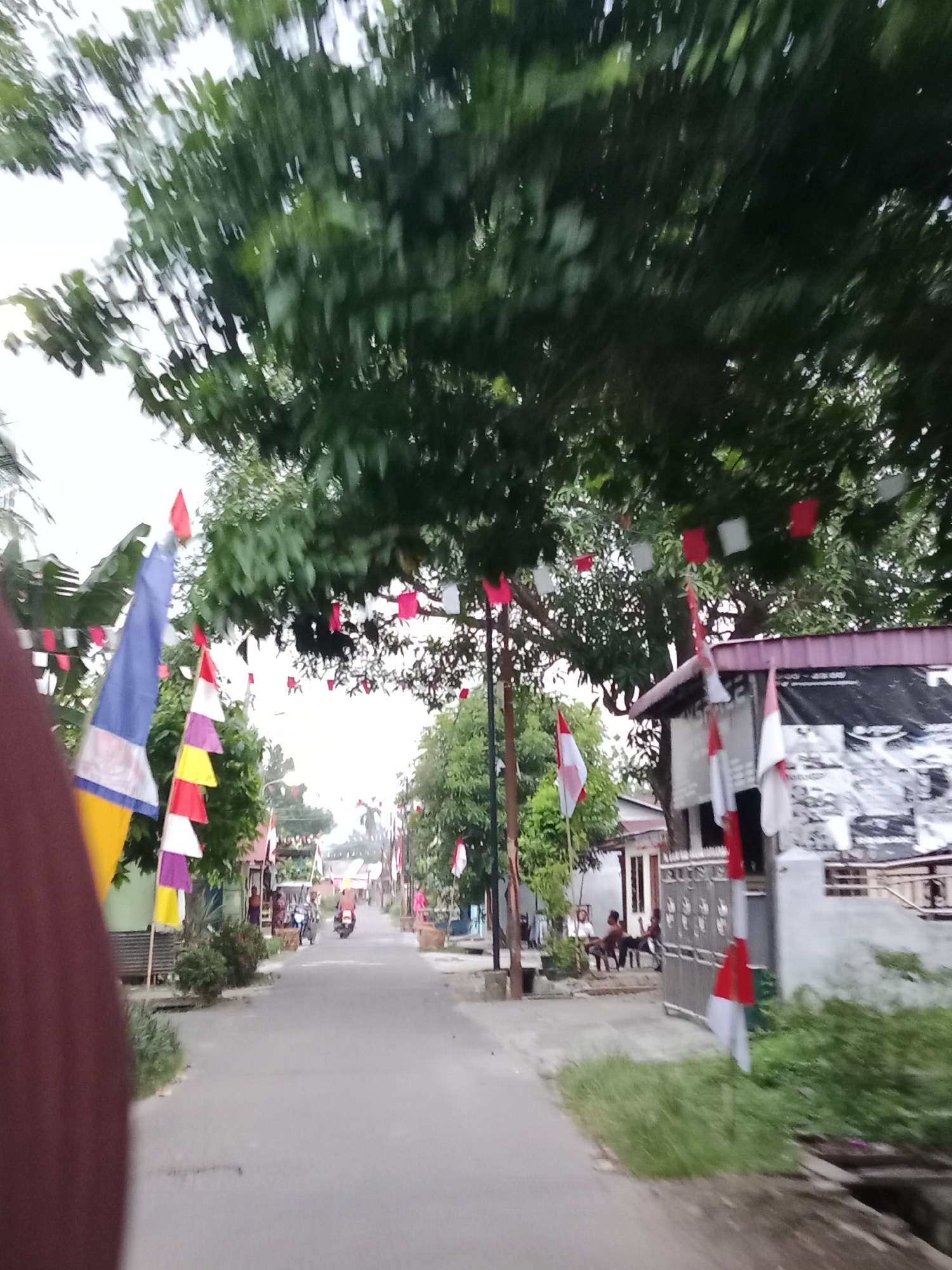 The image shows a street scene in the city of Binjai, Indonesia. The street is lined with trees and houses. There are several Indonesian flags visible, as well as a string of red and white pennant flags hanging from the trees. A few people are seen sitting on a porch on the right side of the street, and a couple of motorcycles are present in the distance. The overall scene suggests a festive or commemorative occasion.