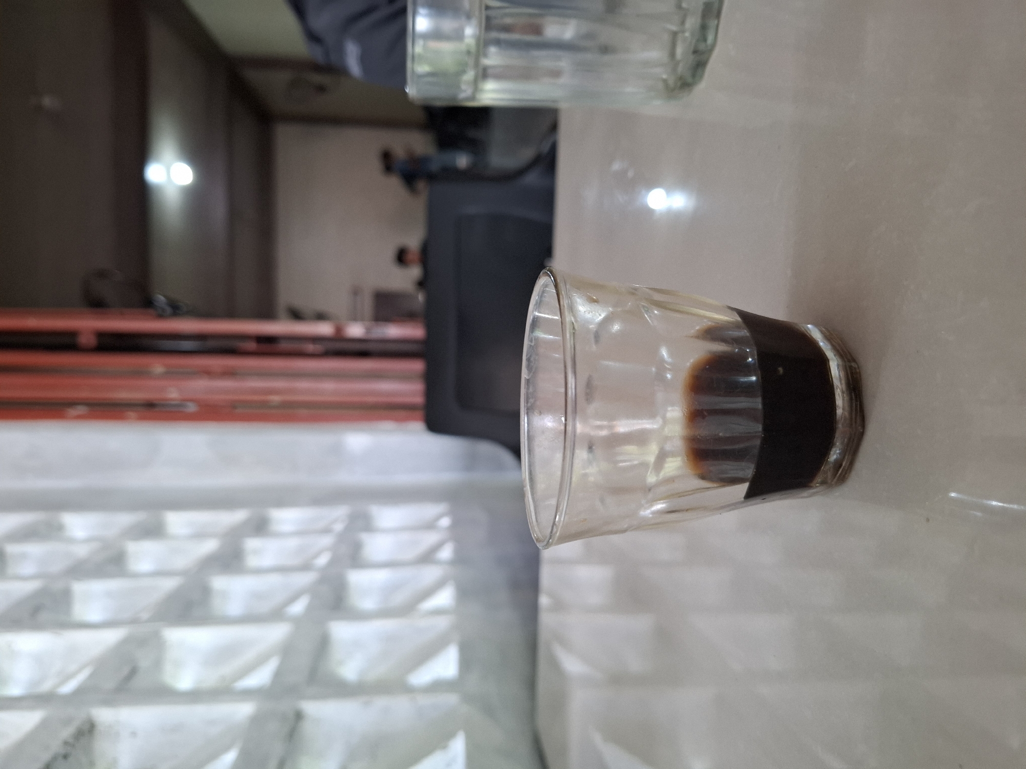 The image, originally rotated, depicts an indoor scene, likely a cafe or eatery, in Lhokseumawe, Indonesia. In the foreground, two clear, faceted glass cups are placed on a light-colored, reflective table surface. The cup closer to the viewer contains a small amount of dark, viscous liquid at its bottom, suggesting it has been mostly consumed, likely coffee or a dark beverage. The second cup, positioned above the first, appears mostly empty or contains clear liquid.
In the mid-ground, a horizontal red railing or barrier runs across the frame. Beyond this, the background is somewhat blurred. To the left, a light-colored wall or partition features a distinctive grid-like pattern, possibly for decoration or ventilation. To the right, two bright, circular light sources are visible against a darker wall, indicating artificial illumination. Faintly visible in the distant background are blurry figures of people and a dark, rectangular object, possibly furniture or equipment. A person wearing a grey long-sleeved shirt or jacket is partially visible in the upper background, near one of the glasses.
The scene suggests a casual setting for relaxation or gathering. No specific activity is ongoing, but the presence of cups implies that beverages have been served or are about to be consumed. The time of day is not discernible, but the reliance on artificial lighting suggests it could be evening or a dimly lit interior. There is no visible text in the image.