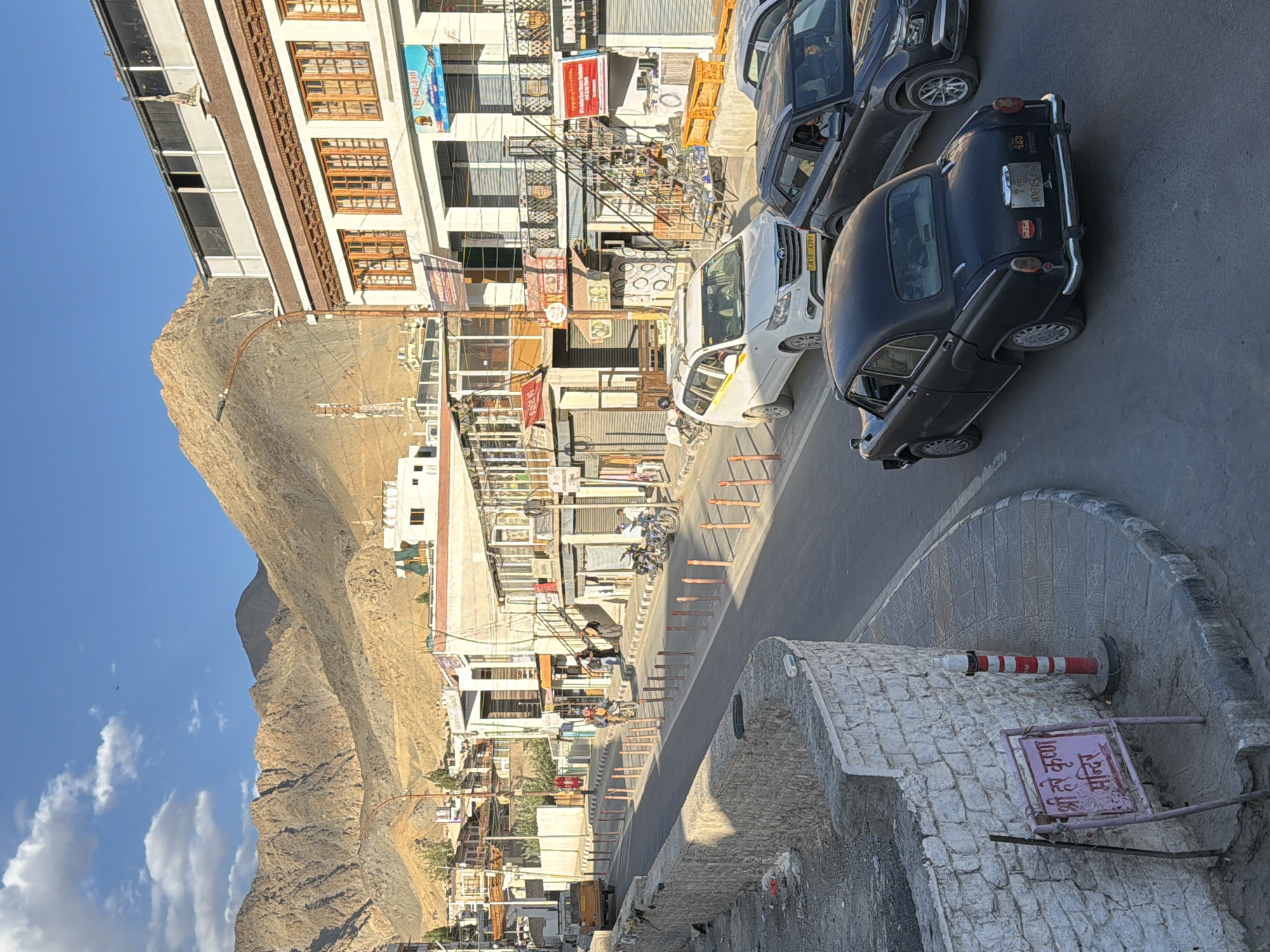 The image shows a street scene in Leh, India. In the foreground, a black car is driving on a paved road. Behind it, a white SUV is also on the road. On the left side of the road, there is a low stone wall. A pink sign with Hindi writing is leaning against the wall. Further down the road, there are several buildings with shops and businesses. The street is lined with orange bollards. In the background, there are steep, arid mountains. The sky is blue with some clouds. A few people are visible on the sidewalk and near the buildings. There are also motorcycles parked on the side of the road. A yellow truck is visible in the distance on the left. The scene appears to be during daylight hours, likely late afternoon given the angle of the sunlight.