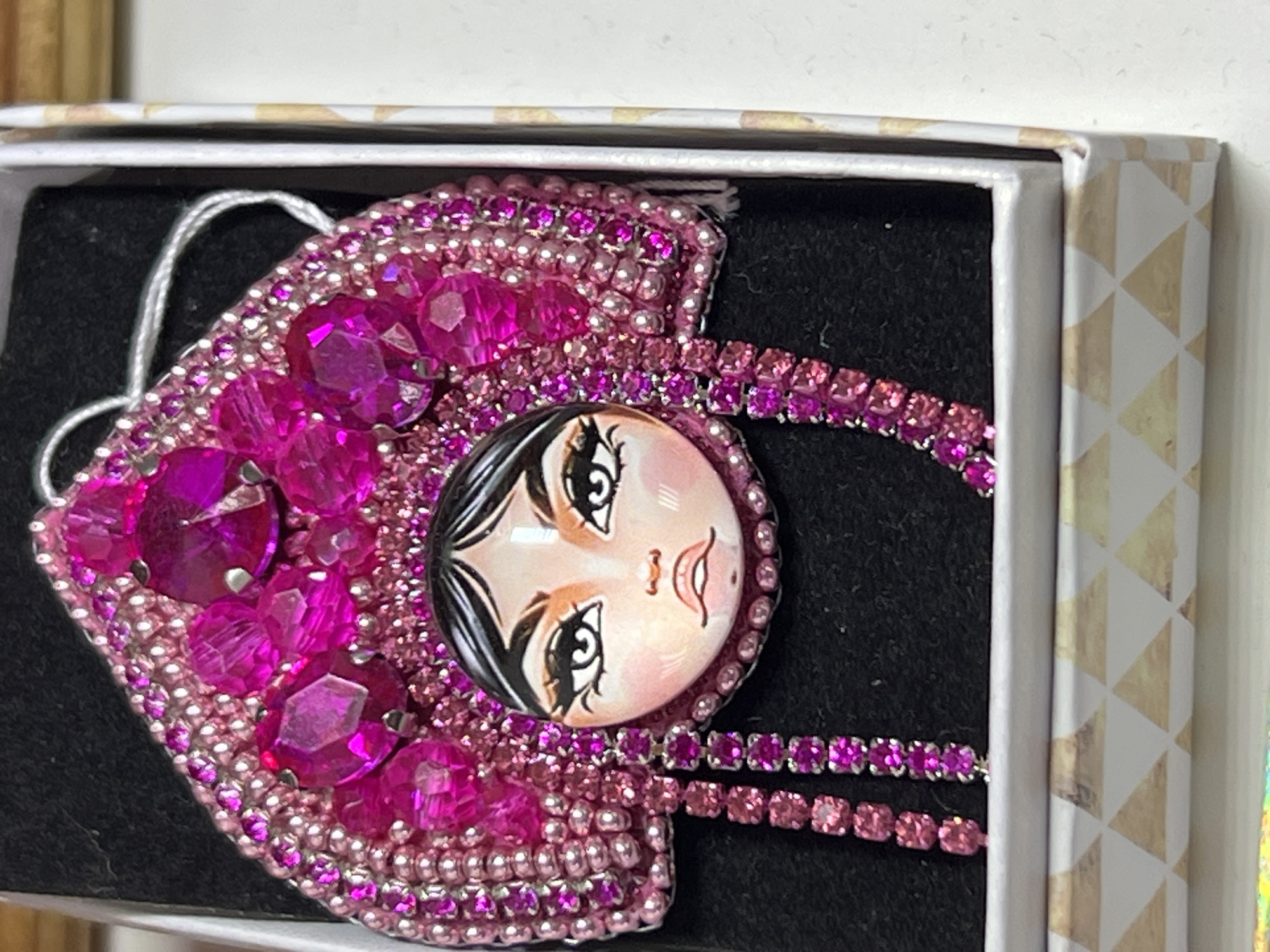 A close-up shot of a handcrafted brooch depicting a woman's face in a jewelry box. The brooch is primarily pink, with various shades and textures created by beads and larger, multifaceted gems. The woman's face is round and has cartoon-like features with large, expressive eyes. The brooch also incorporates rhinestone-like chains. The jewelry box has a patterned edge with gold triangles on a white background. The brooch is set against a black, velvet-like backdrop inside the box. No evidence suggests this image was taken in Chaikovsky, Russia.