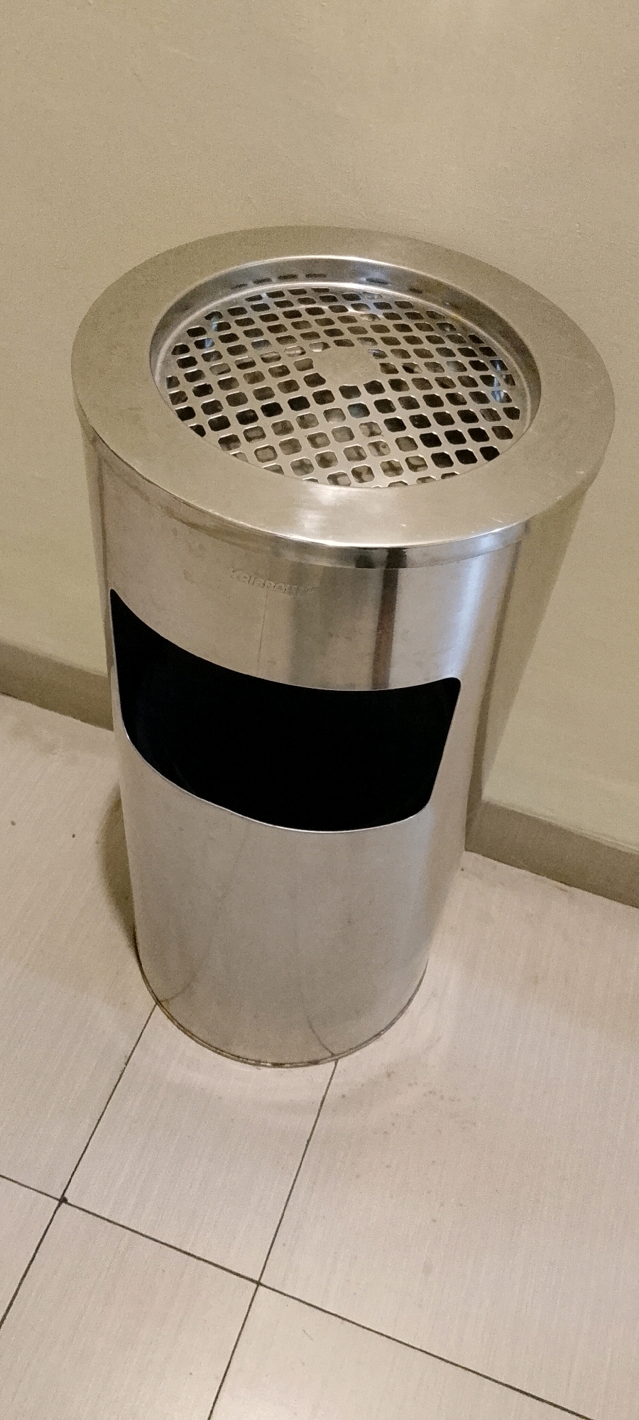 An outdoor ashtray is positioned on a tiled floor in front of a beige wall. The ashtray is made of brushed stainless steel and has a rounded, tapered shape. The top of the ashtray features a metal grate with a pattern of small, square openings, and a slightly raised rim. There is a black, curved cutout on the front of the ashtray, serving as an opening for disposal. Faintly visible on the metal surface above the black cutout is the word "Kalmpons" etched into the steel. The tiled floor is light beige and laid out in a grid pattern with dark grout lines. The wall behind the ashtray is also beige and appears to have a textured finish. The lighting suggests an indoor setting, possibly a public space. The scene is static and shows no people or animals. The photograph is taken from a slightly low angle, looking up at the ashtray.