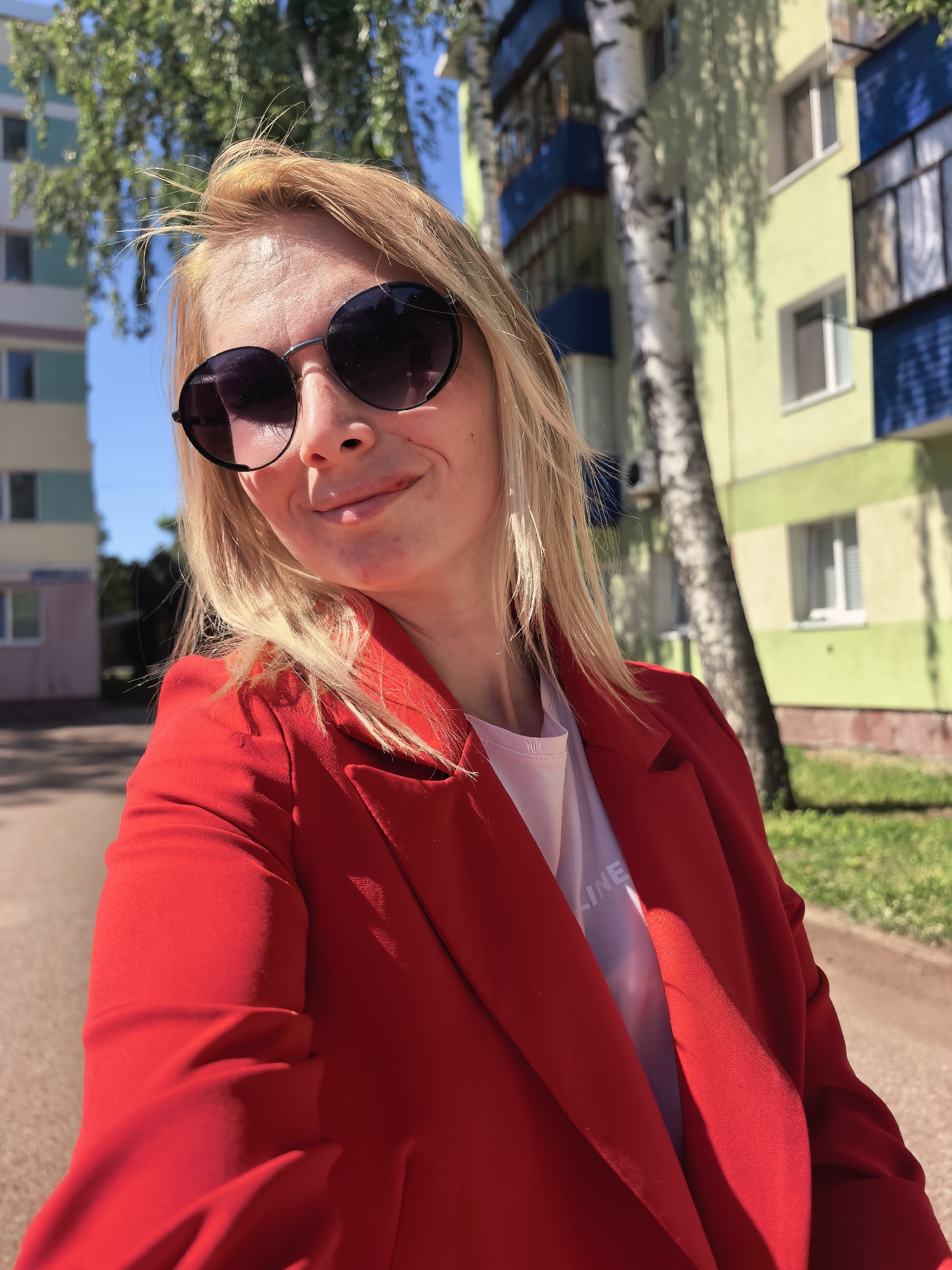 A woman is posing for a selfie outdoors. She is wearing round, dark sunglasses and a bright red blazer over a light-colored t-shirt. Her blonde hair is windblown. The background shows a street and multi-story buildings, one with a green and blue facade and another with light pink walls. A birch tree is visible next to the building with green and blue facade. The location is Sterlitamak, Russia.