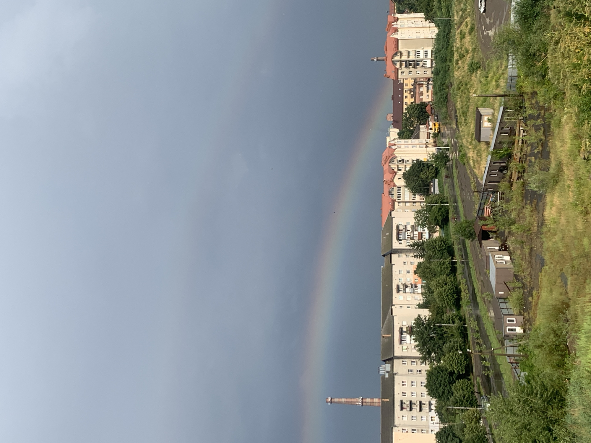 The image shows a wide view of a city in Kolomyia, Ukraine. A rainbow arcs across a dark, cloudy sky. In the foreground, there are several small, weathered sheds and overgrown vegetation. Behind these, rows of apartment buildings with red-tiled roofs are visible, interspersed with trees. A tall, cylindrical industrial chimney stands to the left of the buildings. A yellow bus is parked on a street in the middle ground, and a white car is visible in the far right. The ground in the foreground and middle ground appears wet and muddy, suggesting recent rainfall.