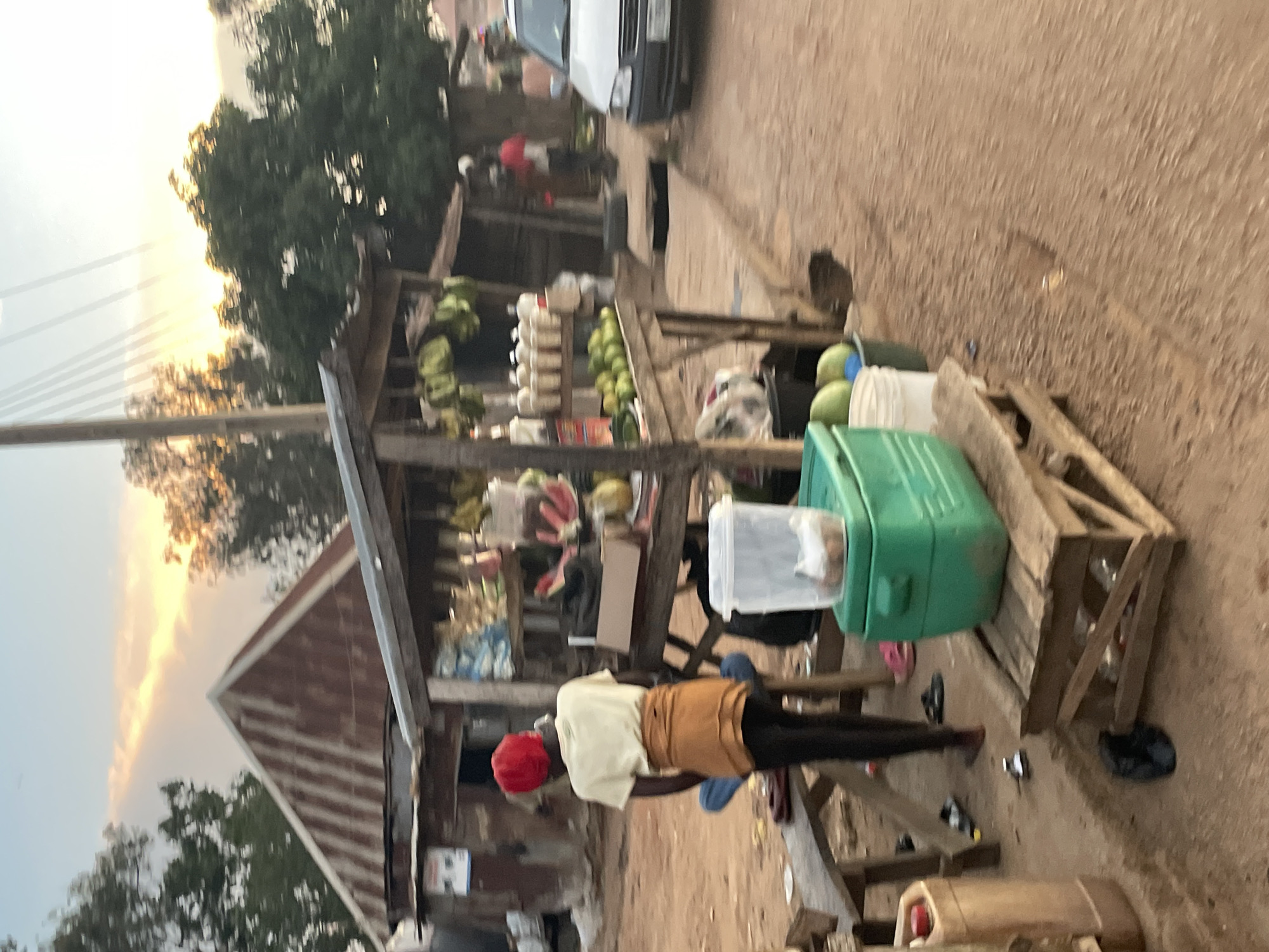 A person wearing a yellow shirt, a brown skirt, and a red headscarf stands facing away from the camera at a roadside market stall in an outdoor setting during what appears to be dusk. The stall is made of wood and displays various items, including fruit like watermelons and green apples, as well as bottled liquids and containers. A green cooler box with a white lid is placed on a wooden crate in the foreground. In the background, a white car is parked on the side of the road, and several buildings with corrugated metal roofs are visible. The ground is unpaved and dusty. The sky shows a warm glow, suggesting either sunrise or sunset.
