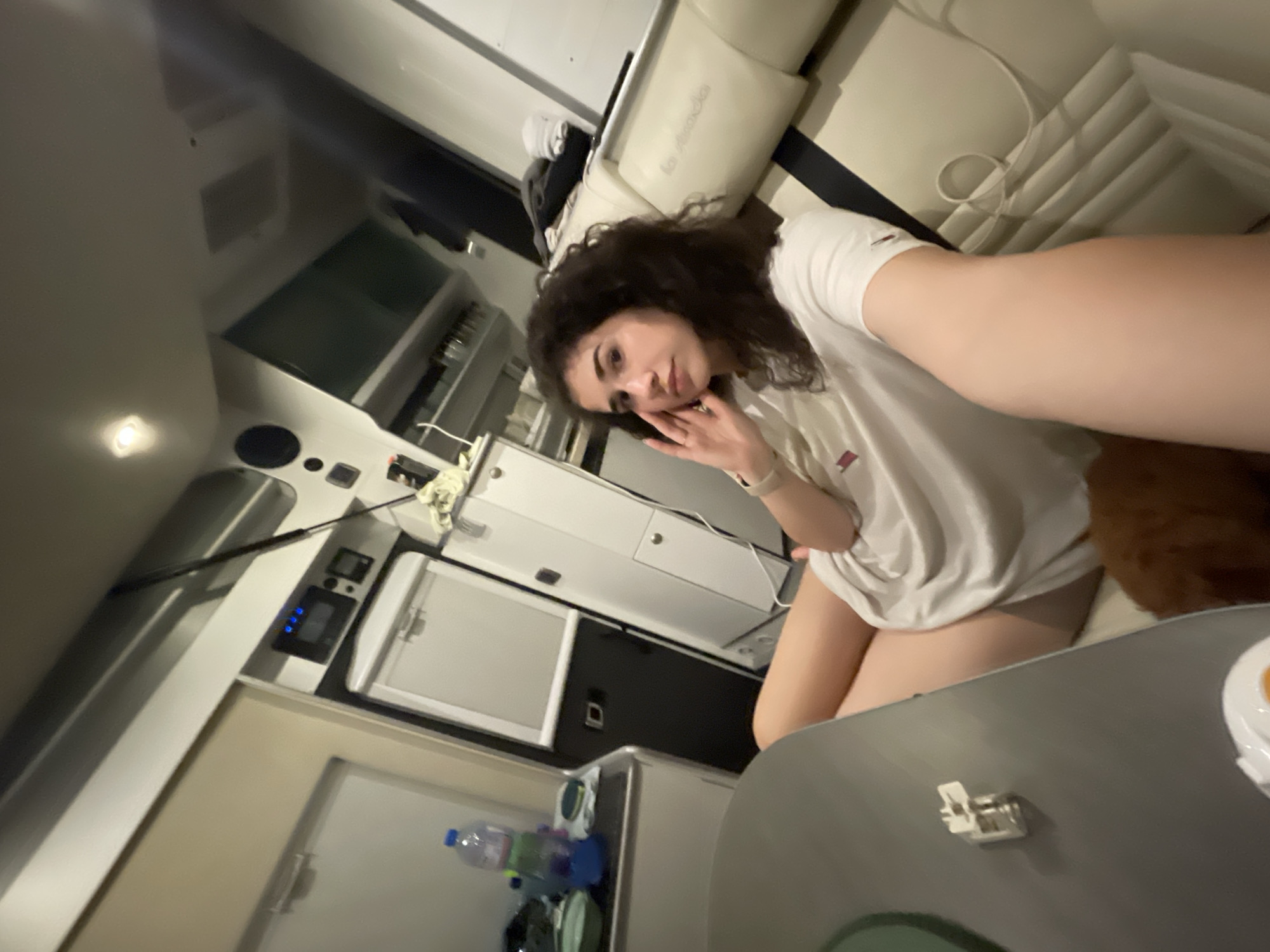 The image displays the interior of a recreational vehicle (RV) or camper van, specified as being in Koprinka, Bulgaria. A young female, exhibiting dark curly hair and wearing a white t-shirt, is seated on a light beige upholstered bench with black accents, featuring the 'la strada' brand logo. She is looking directly at the camera, with her right hand positioned near her face. In the foreground on the left, a dark grey table presents a small metallic component. The vehicle's interior includes a compact white refrigerator/freezer unit, a control panel with blue indicator lights, and overhead storage compartments. A counter adjacent to the refrigerator holds a plastic water bottle, a blue cup, and a green bowl. A portion of a small, brown, furry animal is visible at the bottom of the frame, near the female's legs. The scene is illuminated by ambient interior lighting.