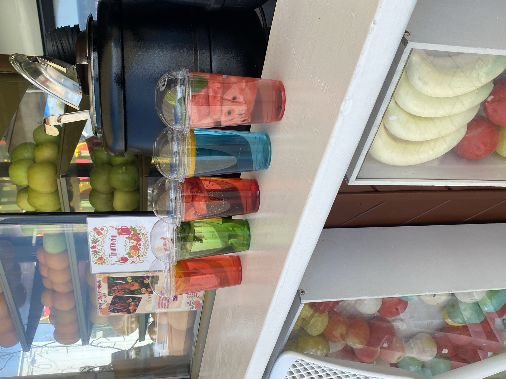 The image shows a refreshment stand, likely located in Sochi, Russia, showcasing a variety of beverages and treats. In the foreground, several clear plastic cups are filled with vibrantly colored drinks, possibly fruit-infused water or tea. The top cup contains pieces of watermelon, while the others display blue, red, green, and orange hues. Behind the drinks are shelves displaying fruit, including green apples and oranges, as well as a menu board with Russian text. A display case containing slices of melon and red fruit is situated to the right of the beverages, with another case showing what appears to be candy. The stand is constructed with a combination of white and brown materials, suggesting an outdoor setting.