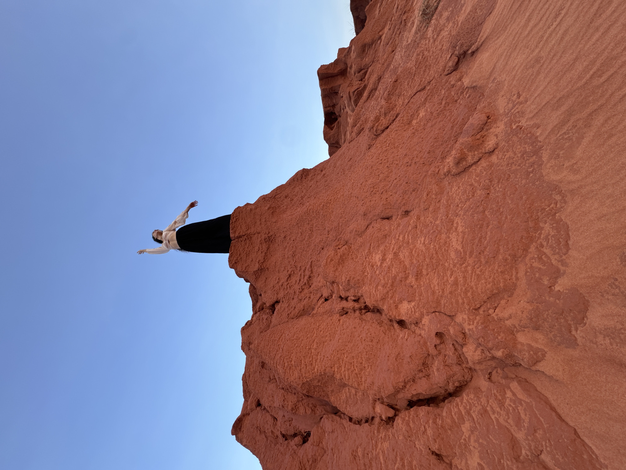An outdoor, low-angle shot captures a person standing triumphantly on the edge of a towering red rock formation against a vast, clear blue sky. The person, appearing to be a woman with dark hair, is positioned on the upper right side of the frame, looking upwards with her arms outstretched and palms open, as if embracing the sky or enjoying the expansive view. She is dressed in a light-colored, long-sleeved top and dark, possibly black, trousers or a skirt.
The prominent red rock formation occupies the right and bottom portions of the image, displaying rich reddish-brown and orange tones. Its surface is rugged, characterized by visible textures, sedimentary layers, and natural cracks, suggesting an arid, desert-like environment. The sky above is a uniform, bright blue, completely cloudless, indicating clear and sunny weather. The lighting is even, suggesting it could be late morning or early afternoon. The overall mood conveys a sense of freedom, peace, and awe in nature. This type of red rock landscape is characteristic of Mui Ne, Vietnam. No visible text is present in the image.