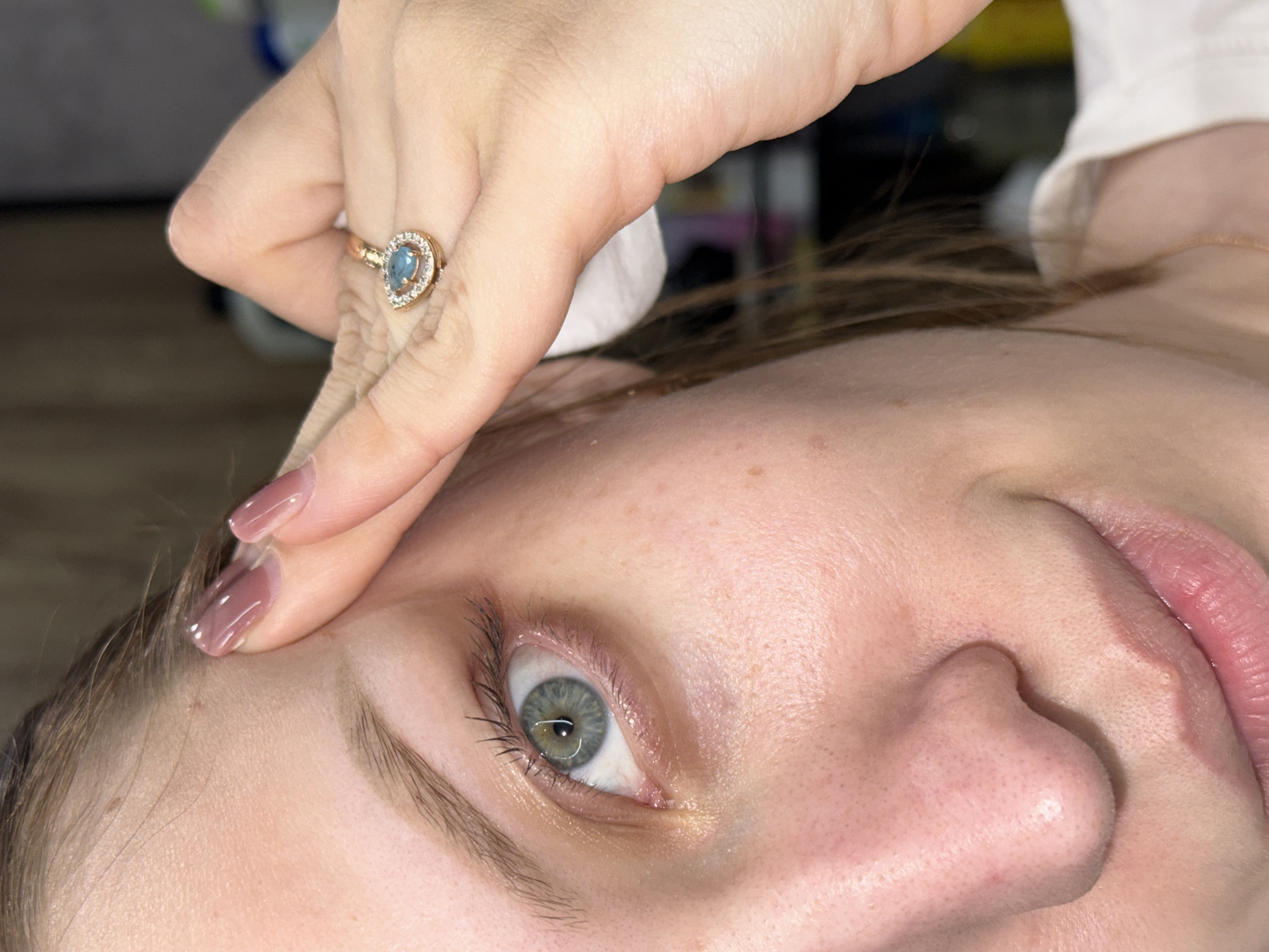 A close-up, tilted shot depicts a person's face and hand. The person has pale skin, light brown hair, and a gray-green eye. A hand, with long fingernails painted a nude color, is touching the person's forehead. The hand has a gold ring with a teardrop-shaped, blue stone surrounded by smaller diamonds. The background is blurred, but suggests a clinical or personal care setting, with a white surface and hints of other items out of focus. The image does not show any elements allowing to determine if it has been taken in the city of Reshetnikovo, Russia.