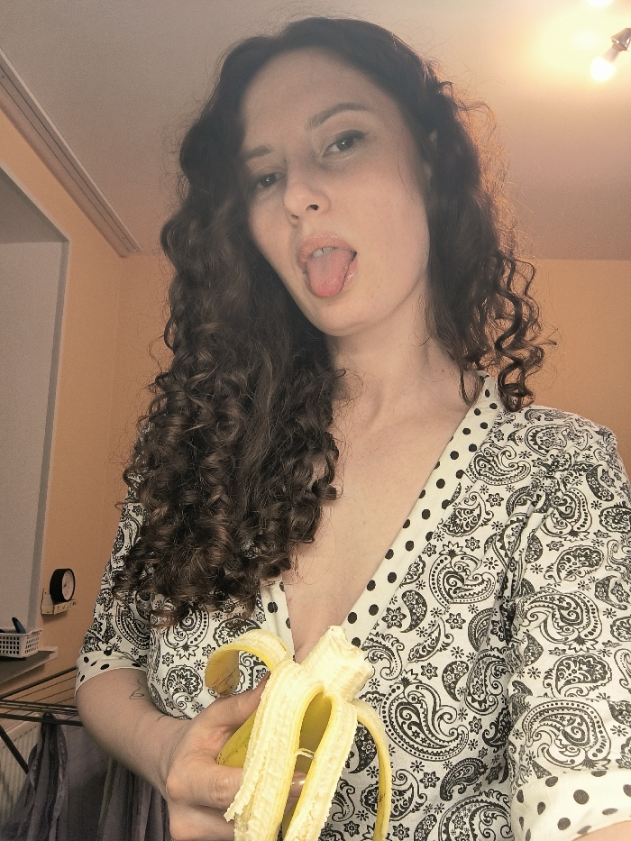 This image depicts an adult female subject in an indoor residential setting in the city of Saint Petersburg, Russia. The woman, who has long, dark, curly hair, is positioned centrally, looking directly at the camera with her tongue extended from her mouth. She wears a v-neck top with a white background adorned with a black paisley pattern and black polka dot trim along the neckline. A ring is visible on the ring finger of her left hand. She holds a partially peeled ripe yellow banana in her left hand; the top portion of the fruit is exposed, while the lower peel segments remain attached. The background consists of a light peach-colored wall on the left and a white ceiling. Two exposed light bulbs, one of which is illuminated, are visible on the ceiling in the upper right. On the peach wall to the far left, a dark, circular object resembling a wall clock is mounted, below which a white plastic mesh basket rests on a surface. Further down, a portion of a metal drying rack with dark fabric hanging on it is visible.