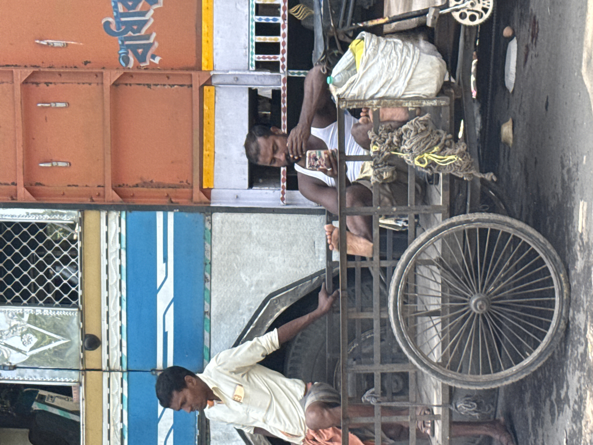 The image shows two men in Kolkata, India. One man is sitting on a rickshaw and looking at his phone. The other man is pushing the rickshaw. The background includes the side of a truck or bus.