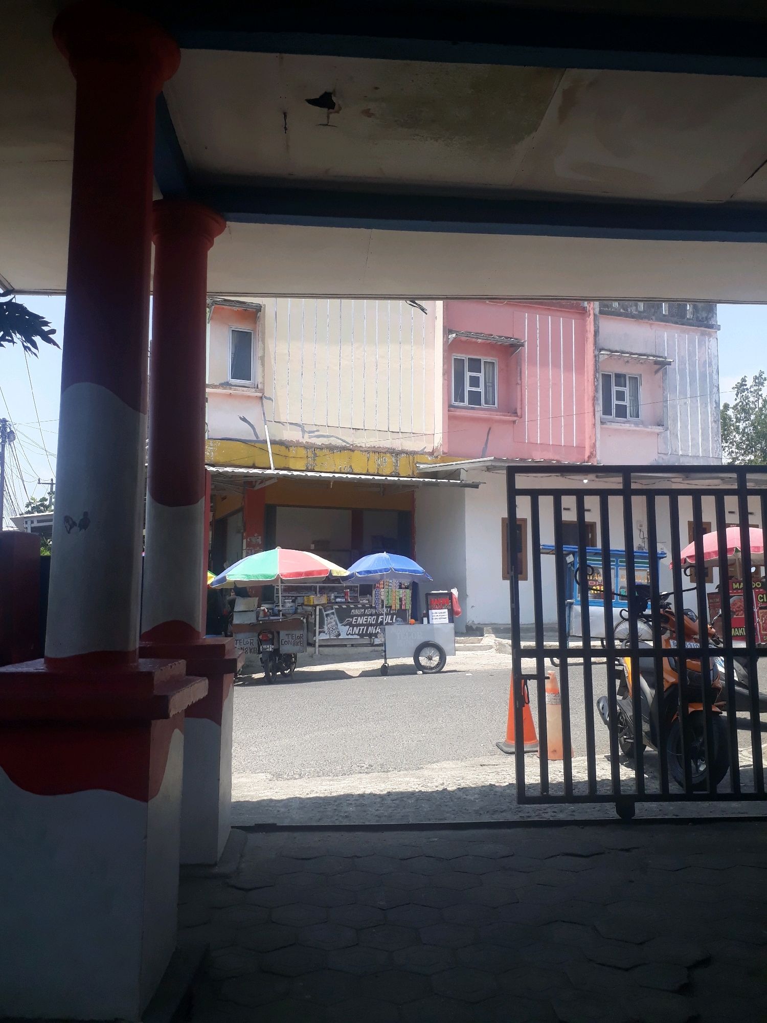 The image shows a street scene in Pangkalpinang, Indonesia, viewed from under an overhang supported by two red and white columns. In the middle ground, two food carts are visible on the street. The cart on the left has a colorful umbrella and is selling various items. The cart on the right also has an umbrella and appears to be selling food. Behind the carts, a row of multi-story buildings with light-colored facades and pink accents lines the street. A black metal fence is in the foreground on the right, obscuring a parked orange motorcycle. Traffic cones are also visible near the fence. The sky is clear and blue. The overall impression is of a typical urban environment with small businesses operating along the street.