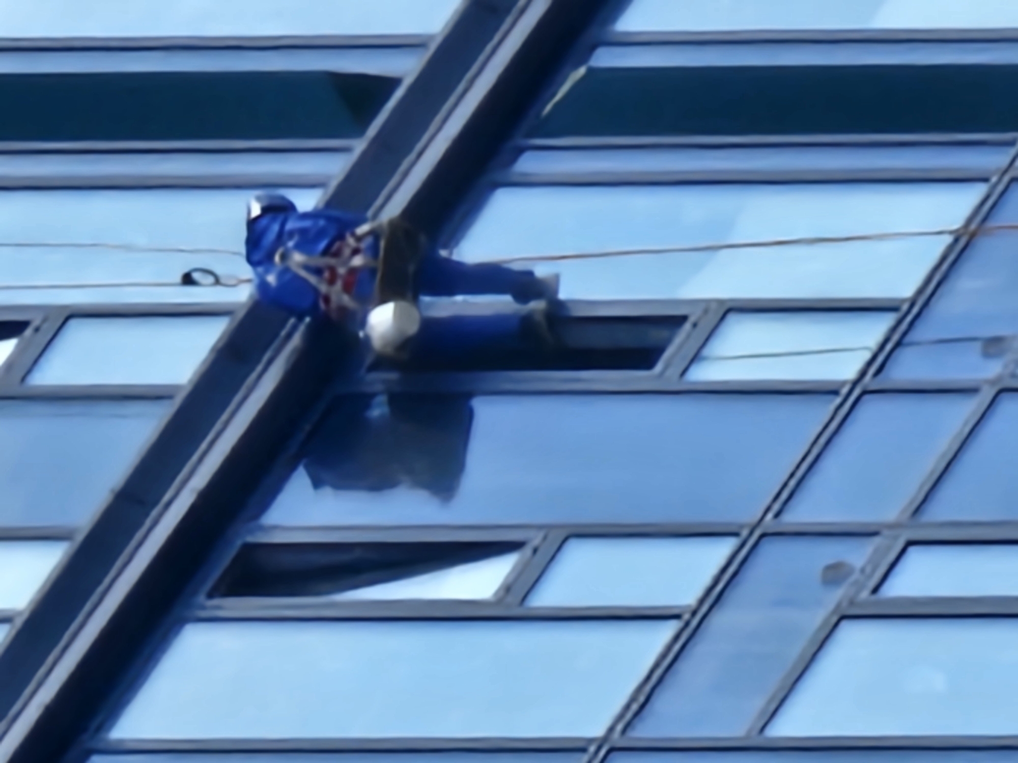 A person wearing a blue jumpsuit and safety harness is suspended by ropes on the side of a modern glass-covered building, likely engaged in window cleaning or building maintenance. The building's facade is composed of large, rectangular blue glass panels framed by dark grey mullions. The scene suggests an urban environment, and the style of the architecture is consistent with contemporary cityscapes. There is no specific visual information in the image to indicate the location as Moscow, Russia.